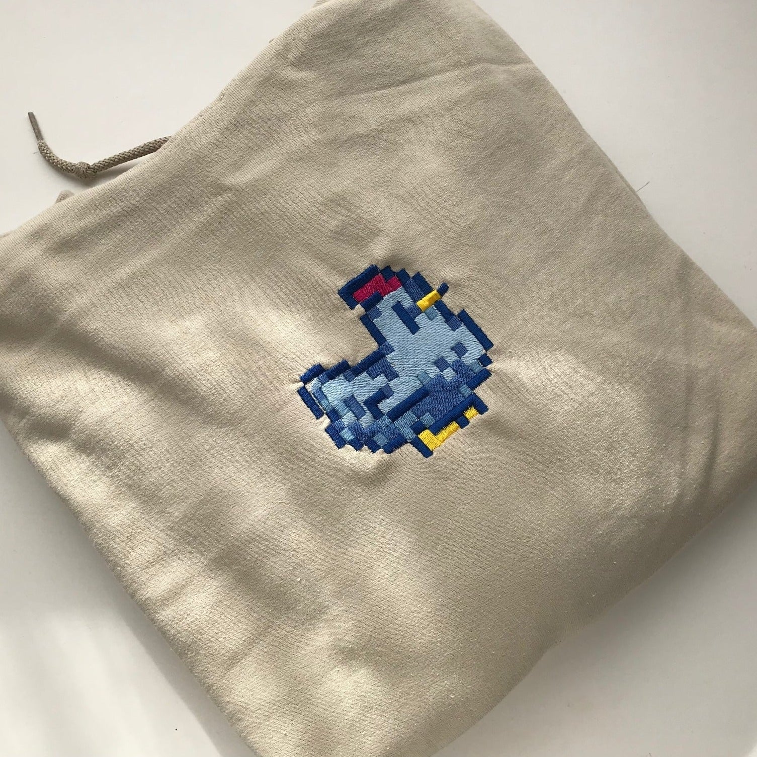 The fabric bag, unisex in style, is beige with a drawstring and showcases an embroidered pixelated design of a blue and yellow bird symbol. It is presented laying flat on a crisp white background and resembles the Stardew Blue Chicken Hoodie by petimint.