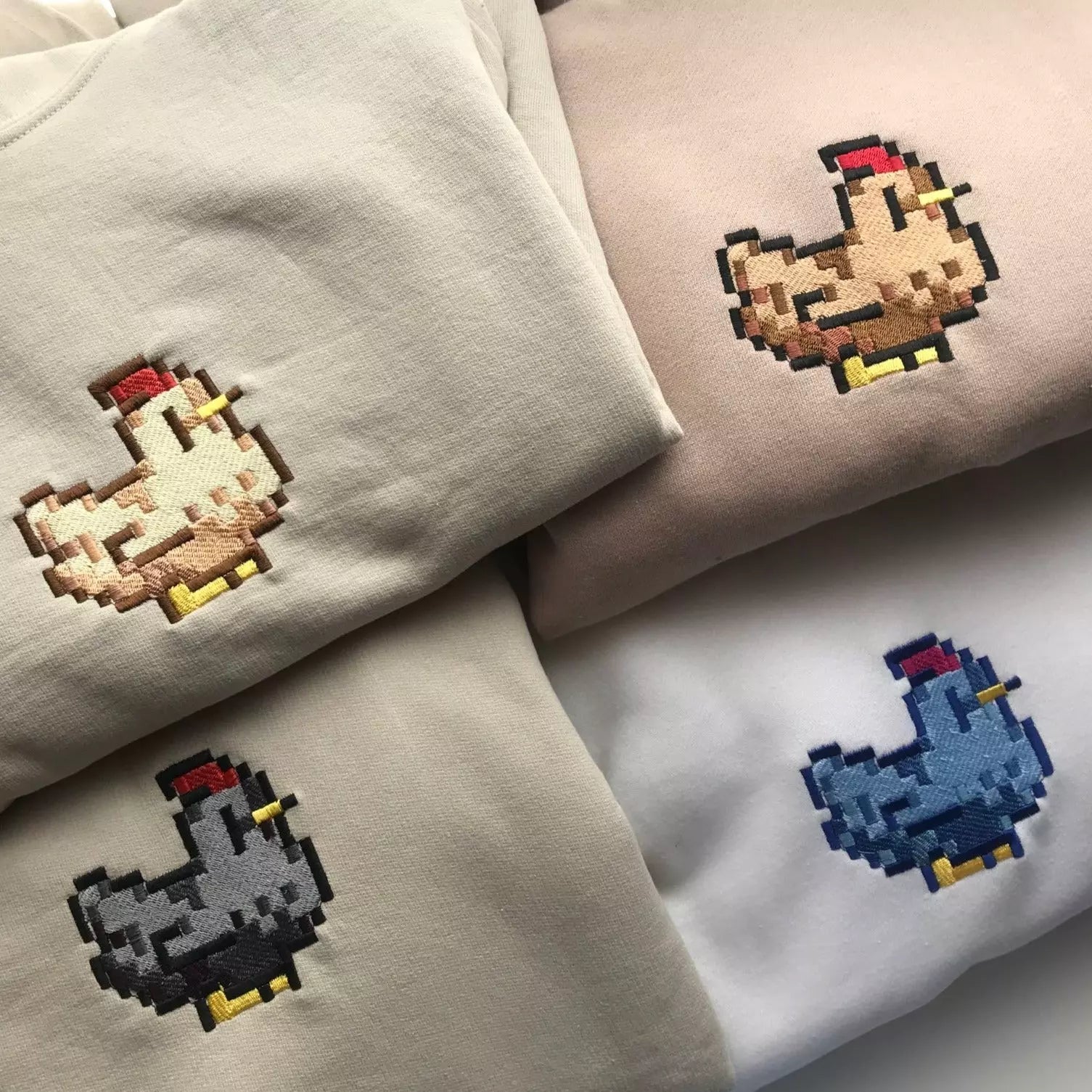 Four folded crewneck shirts in light beige, tan, grey, and white colors feature an adorable pixel art chicken embroidered on the chest. This cozy gamer apparel by petimint includes chickens in colors that match each Stardew Brown Chicken Crewneck: a light chicken on beige, a tan chicken on tan, a grey chicken on grey, and a blue chicken on white.