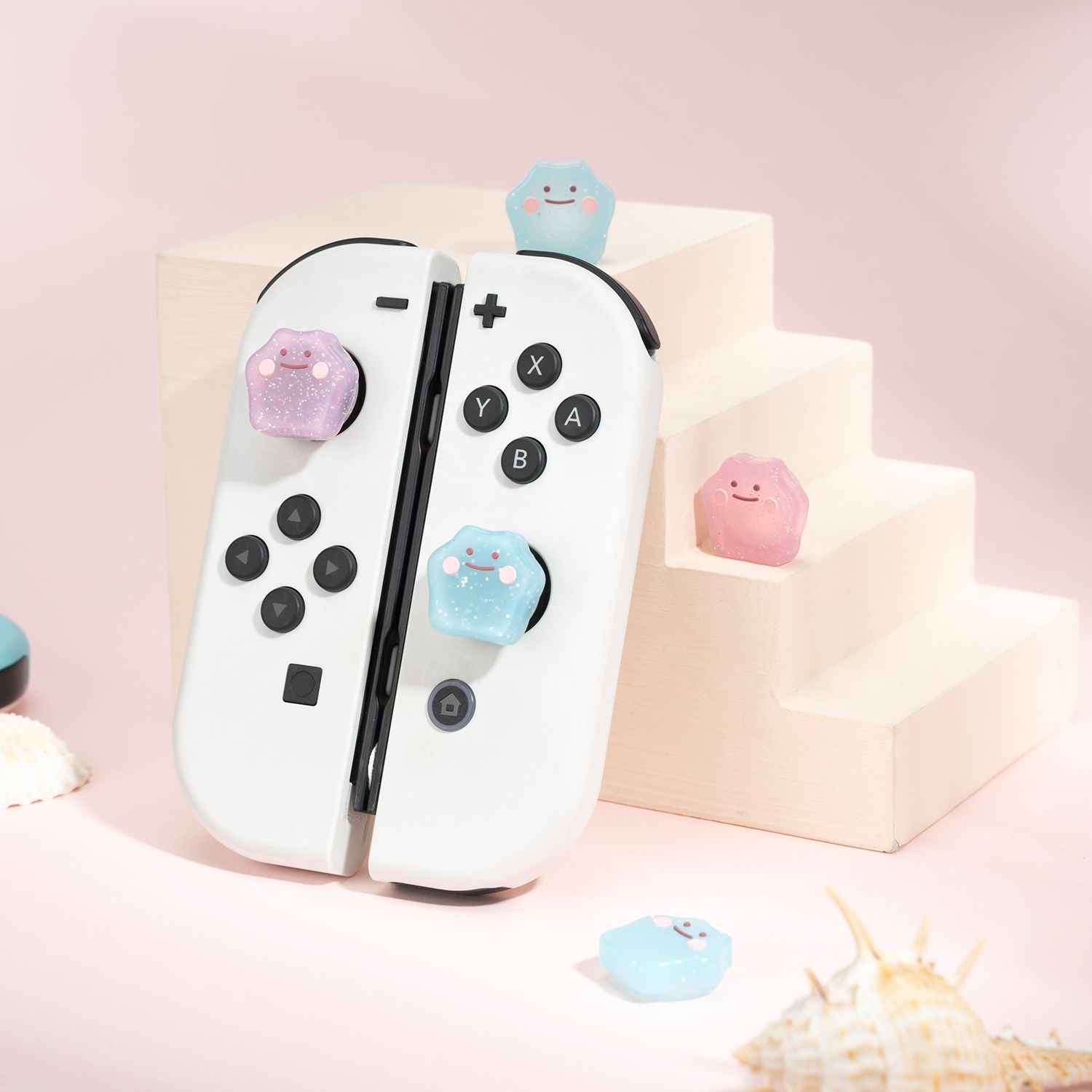 A pair of white gaming controllers with black buttons stand upright against a beige, step-like structure. Small, pastel-colored Glitter Ditto Thumb Grips for Nintendo Switch, featuring smiling faces, rest on the joysticks and steps. Seashells are scattered around the scene, adding a whimsical touch reminiscent of a playful Nintendo Switch setup by petimint.