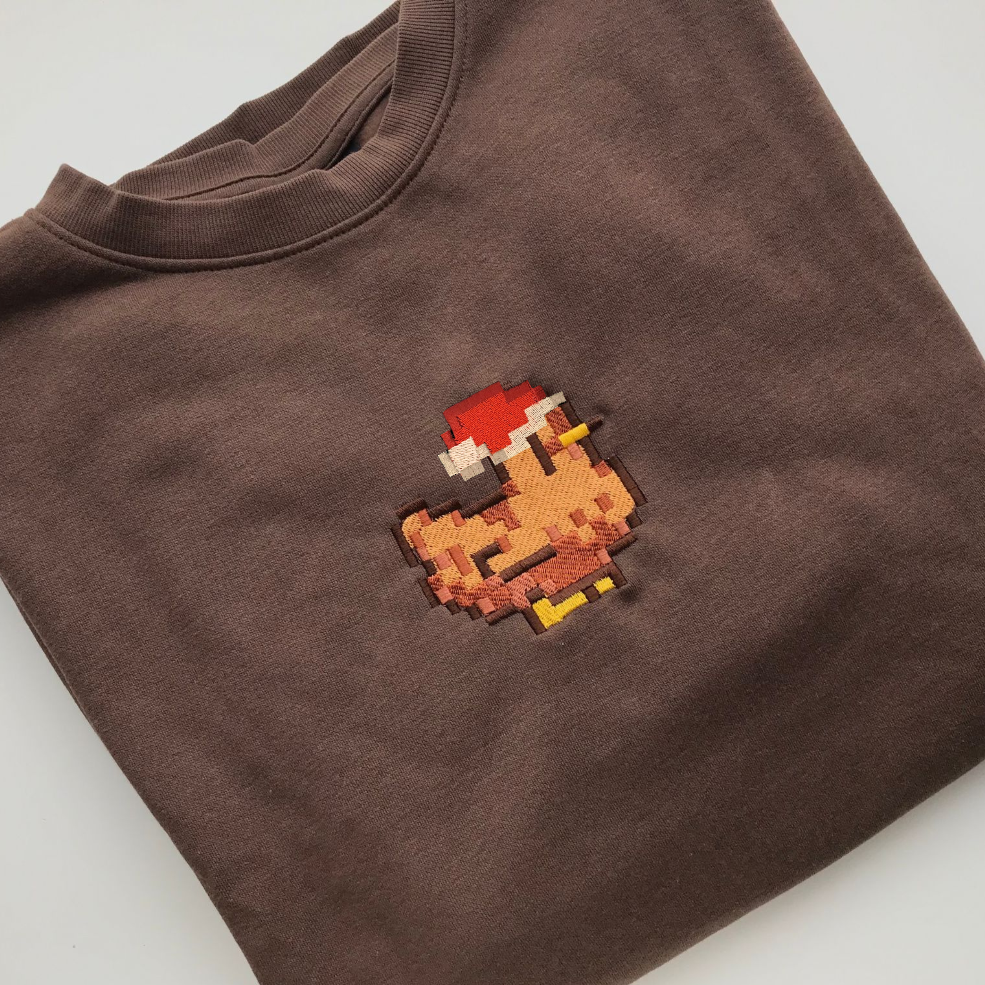 The petimint Christmas Stardew Brown Chicken Sweatshirt features an embroidered design of a pixelated chicken wearing a red hat on the front, making it perfect for any gaming enthusiast. The dark brown sweatshirt is folded and displayed on a white background.