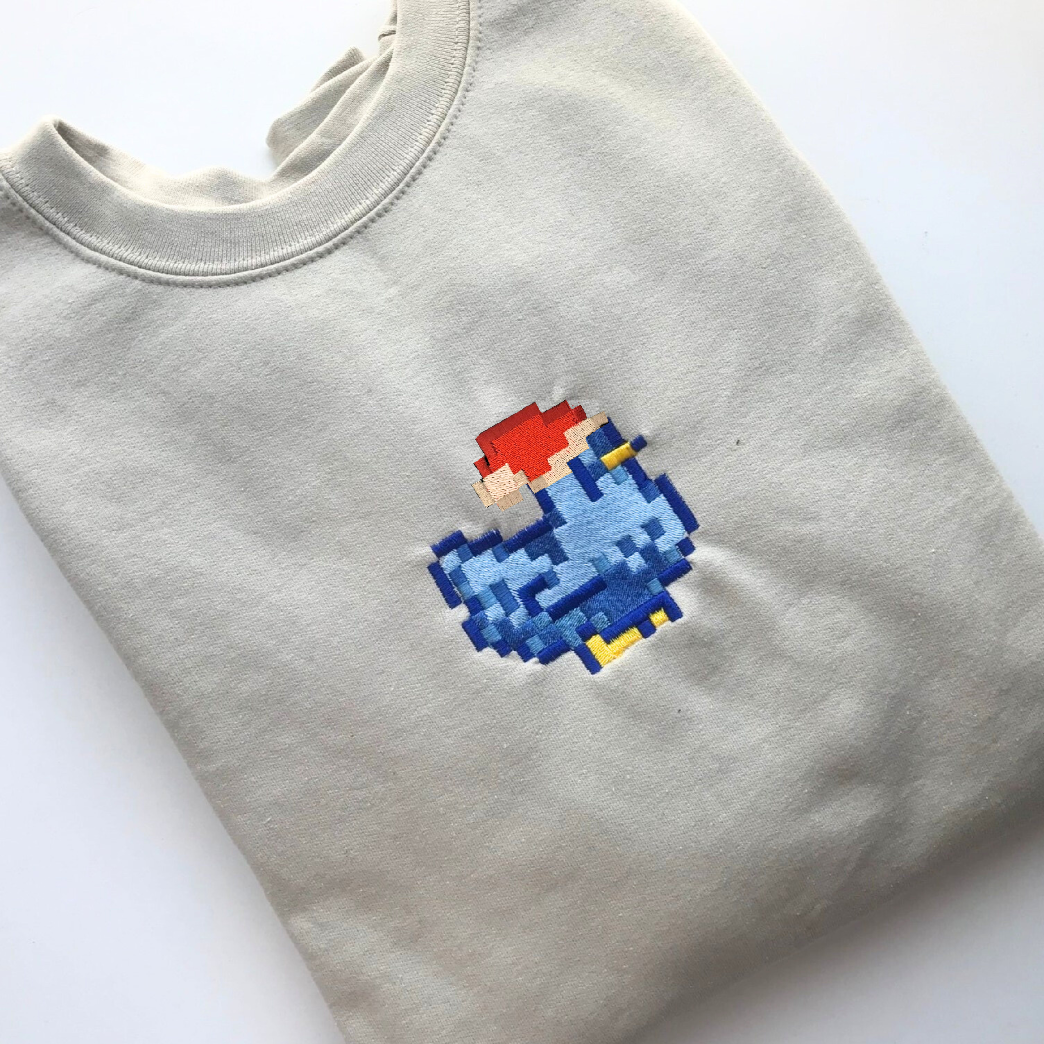 The Christmas Stardew Blue Chicken Sweatshirt by petimint is a folded beige sweatshirt featuring a pixelated embroidered design on the front. The embroidery showcases a character with red hair and a blue outfit, reminiscent of retro video game art. Perfect cozygamer apparel, this sweatshirt is placed on a white surface.