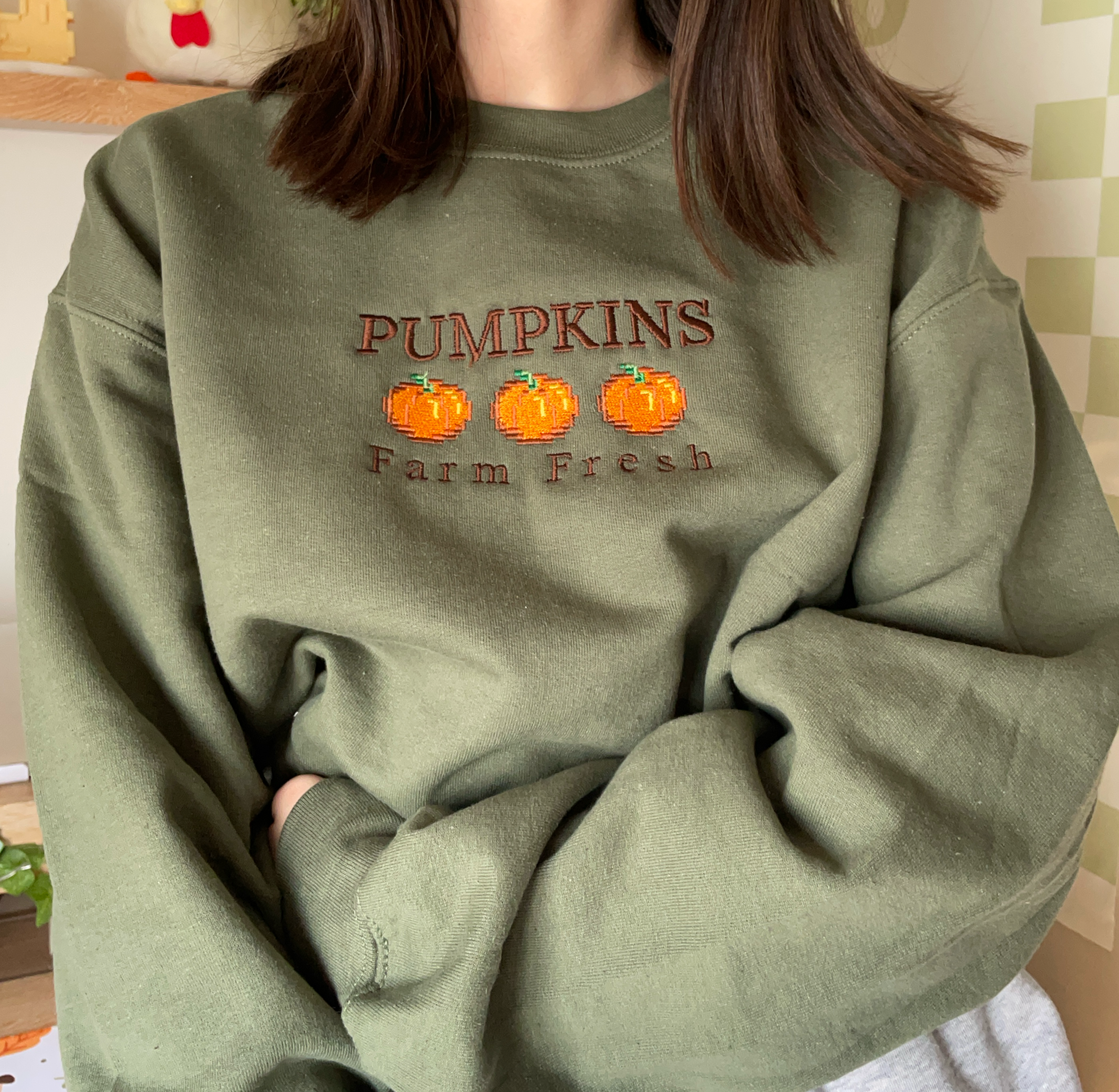 A person is dressed in a petimint's "Pumpkins Farm Fresh Crewneck Halloween Apparel," featuring embroidered pumpkins and the text "Pumpkins Farm Fresh" on the chest. The cozy gamer, with long brown hair, has their arms crossed. The background includes a small potted plant and a checkered wall, creating an ideal atmosphere for a snug gaming setup.