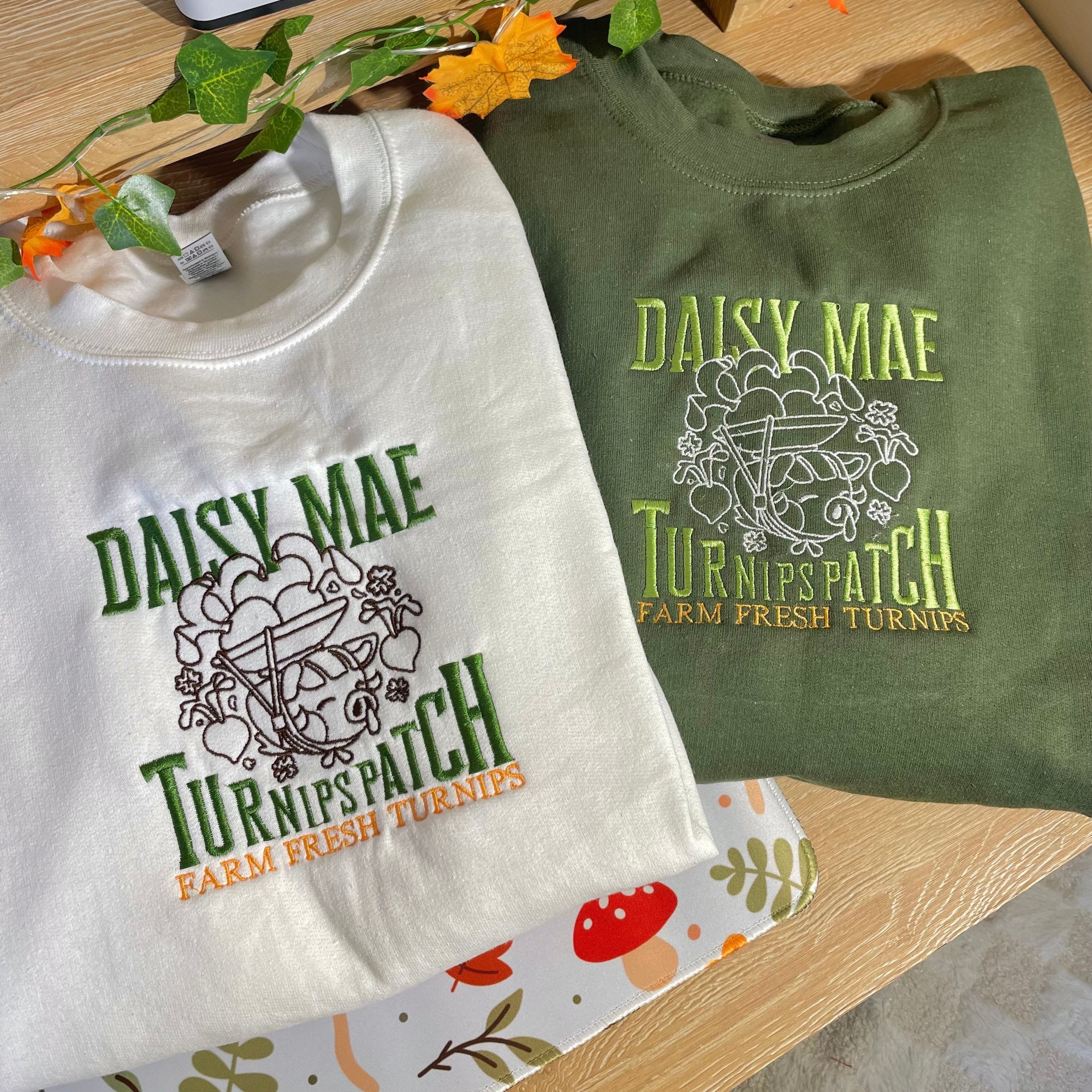 Two sweatshirts from petimint's Turnips Patch Crewneck Gamer Apparel collection are lying on a wooden surface. The white sweatshirt features green and yellow embroidered text reading "Daisy Mae's Turnip Patch," while the green sweatshirt showcases the same design in white and yellow. Above, fall decorations are partially visible, making these perfect additions to any cozygamer's apparel collection.