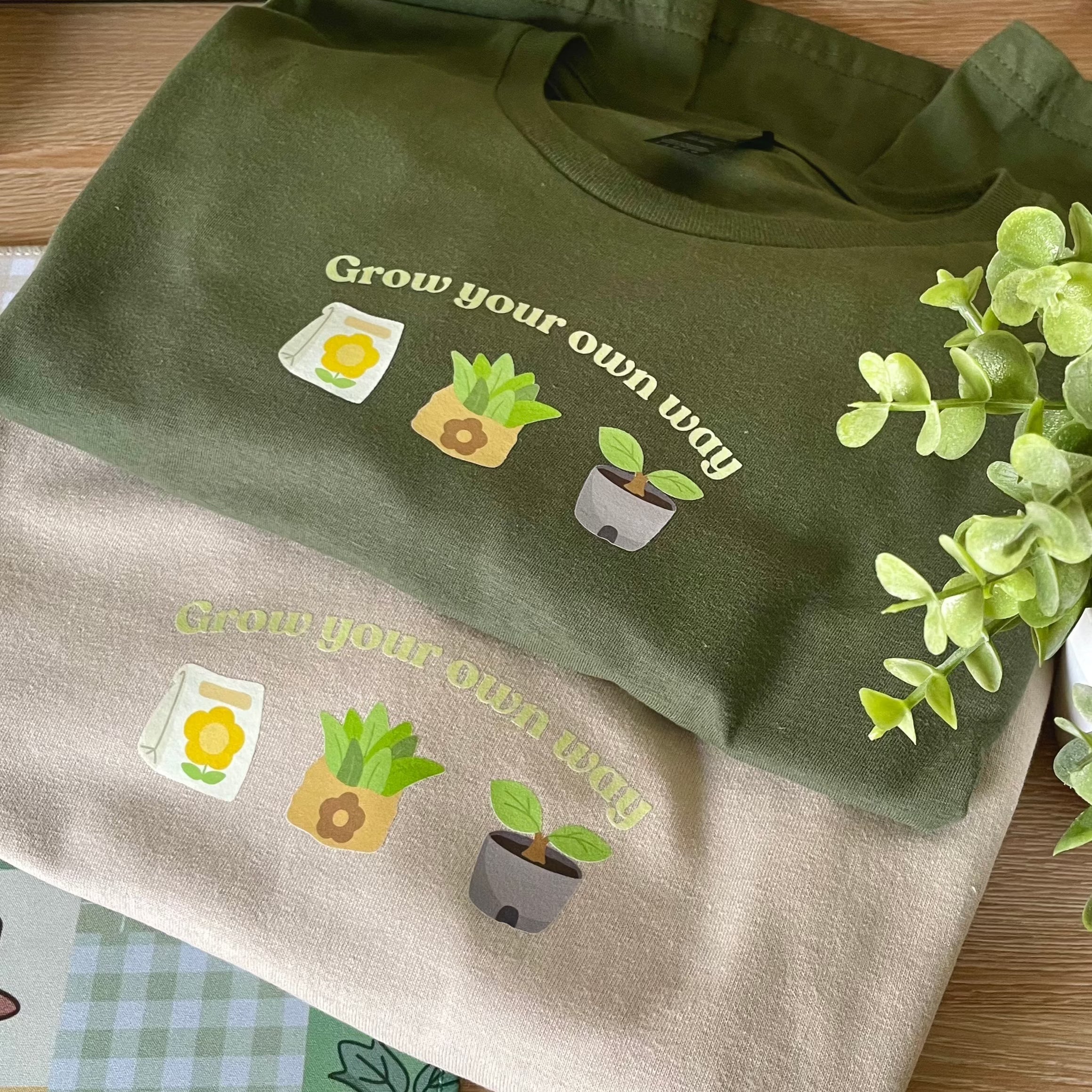 Two Grow your own way ACNH T-Shirts from petimint, one in dark green and the other in tan, both adorned with the phrase 