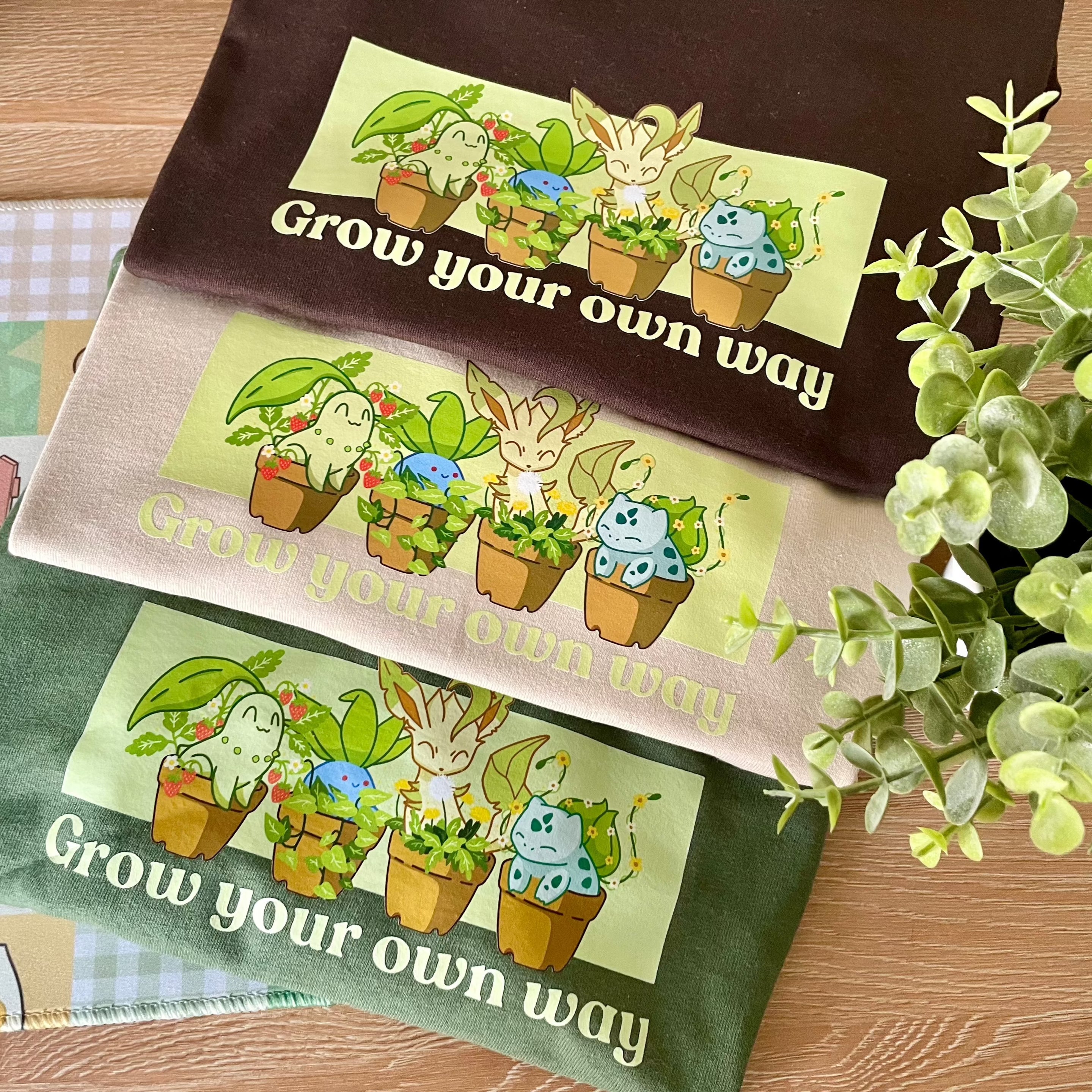 Displayed on a wooden surface next to a small leafy plant are three folded Poképots T-Shirts from petimint in brown, beige, and green. Each shirt features an illustration of cute plant creatures in pots accompanied by the text 