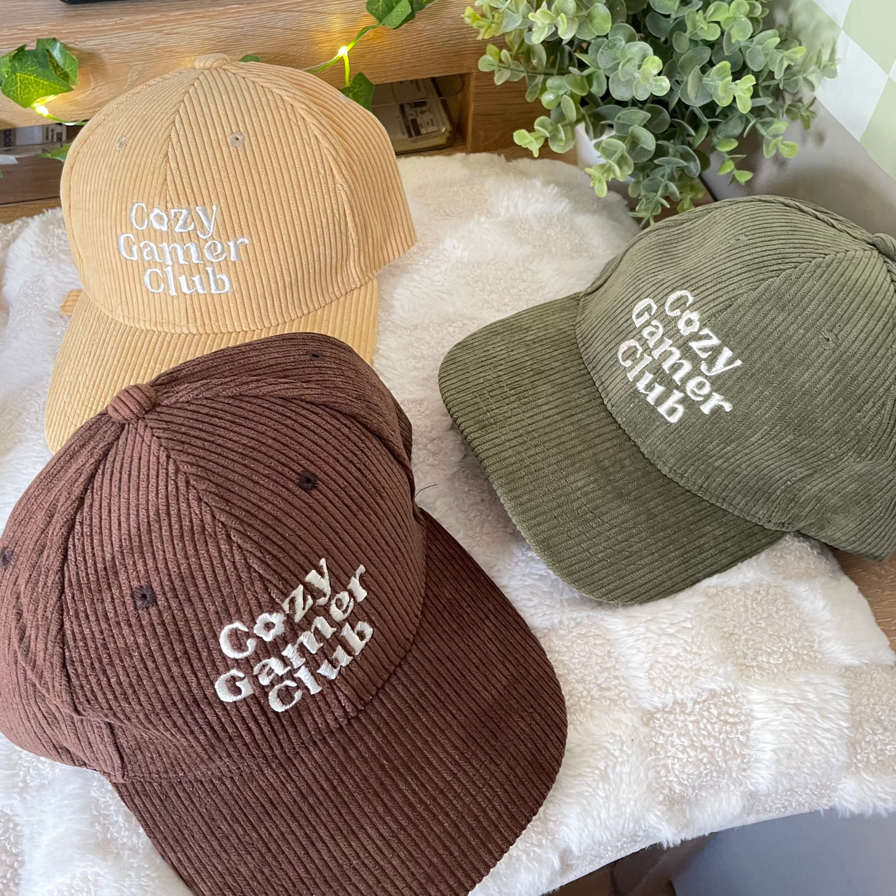 Three Cozy Gamer Club caps by Petimint, featuring "Cozy Gamer Club" embroidered on the front, are displayed on a soft, white surface. The caps come in tan, brown, and green colors. In the background, green plants and twinkling lights add a touch of Petimint charm.