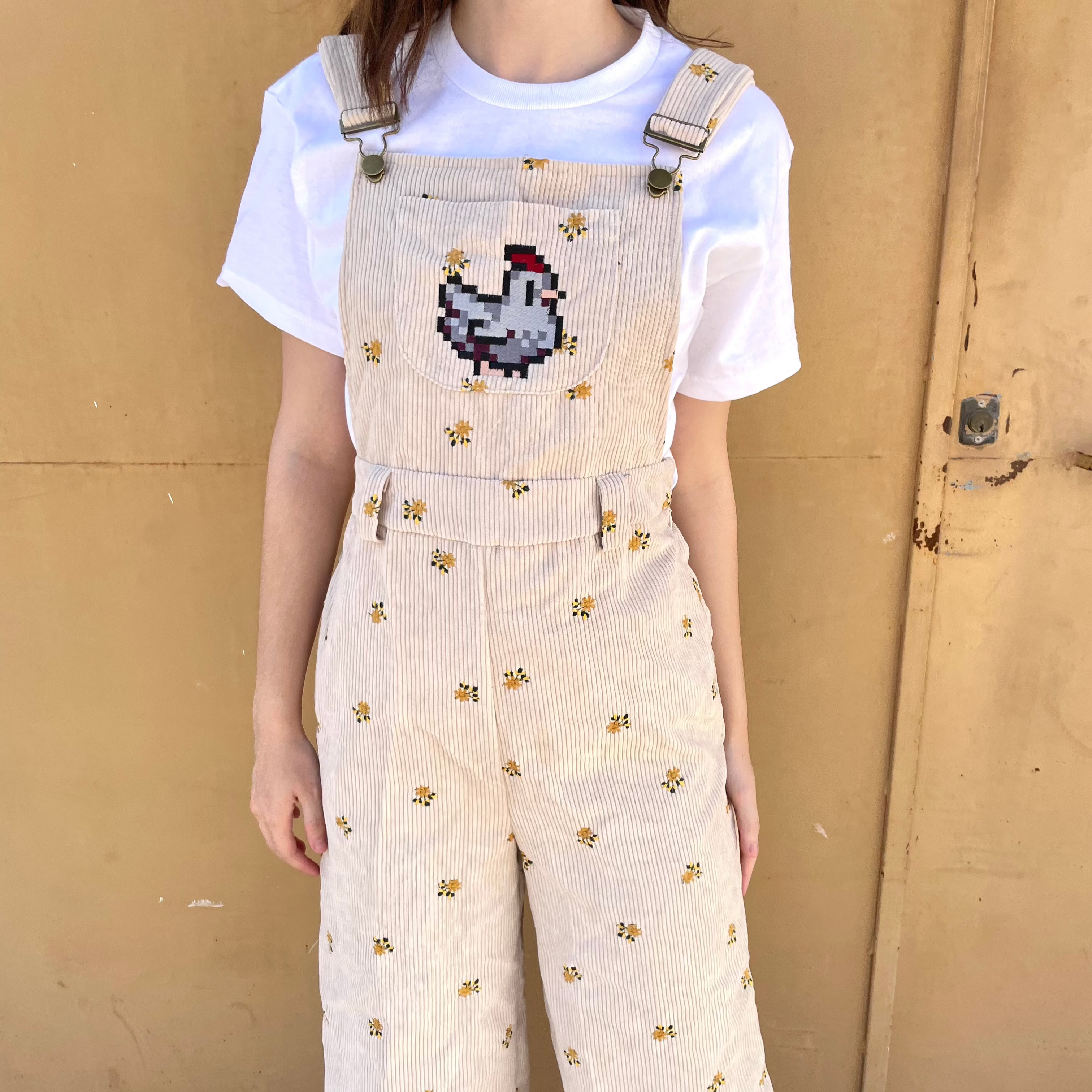 A person is standing against a beige wall wearing the petimint Stardew Chicken Floral Overalls Long Pants, featuring a small yellow floral pattern. A pixelated chicken, inspired by Stardew Valley, is prominently embroidered on the front of the overalls. They are also wearing a plain white t-shirt underneath.
