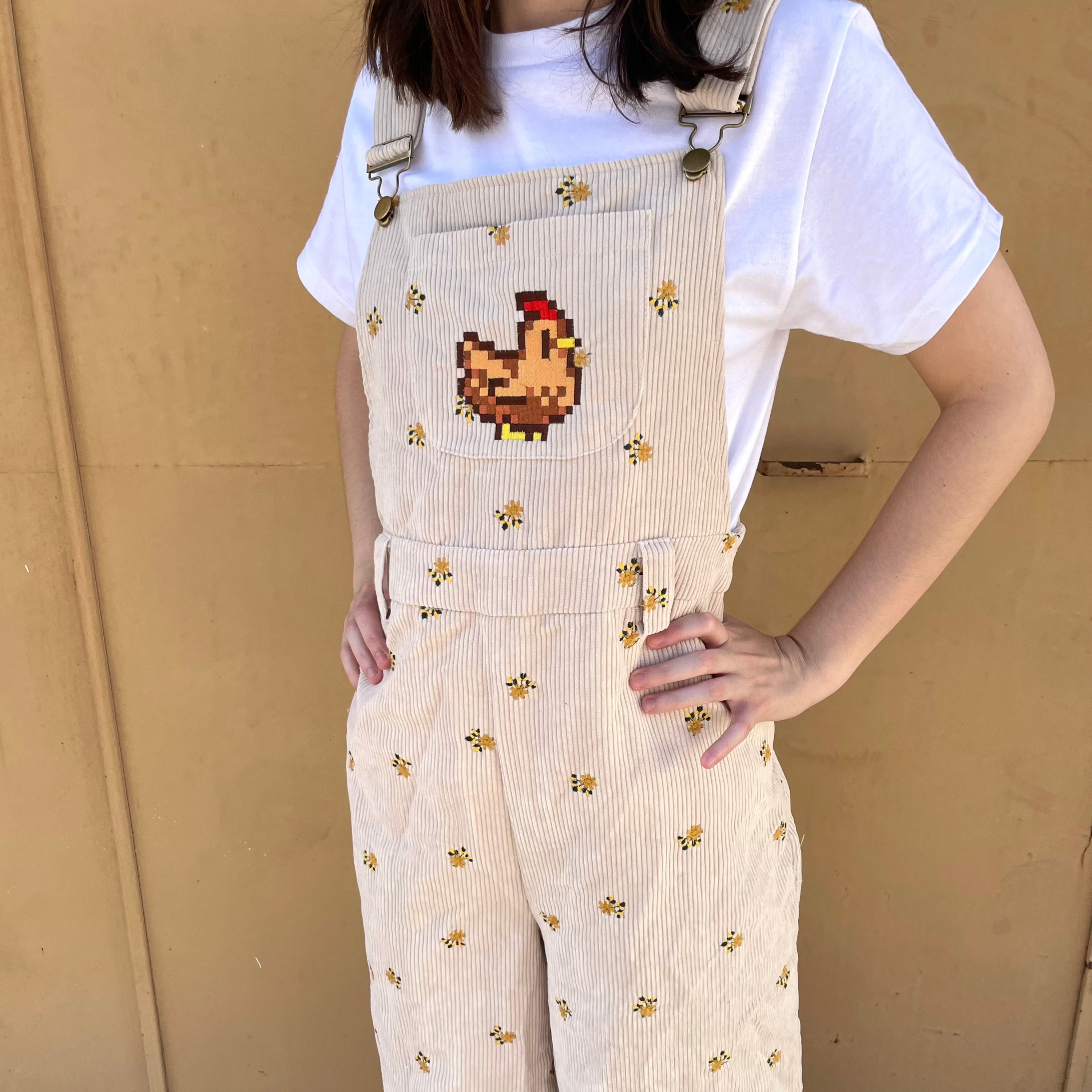 A cozygamer vibe radiates from the individual dressed in a white T-shirt and petimint's Stardew Chicken Floral Overalls Long Pants, featuring small embroidered flowers and a pixelated chicken patch on the front pocket, as they stand against a plain beige background.