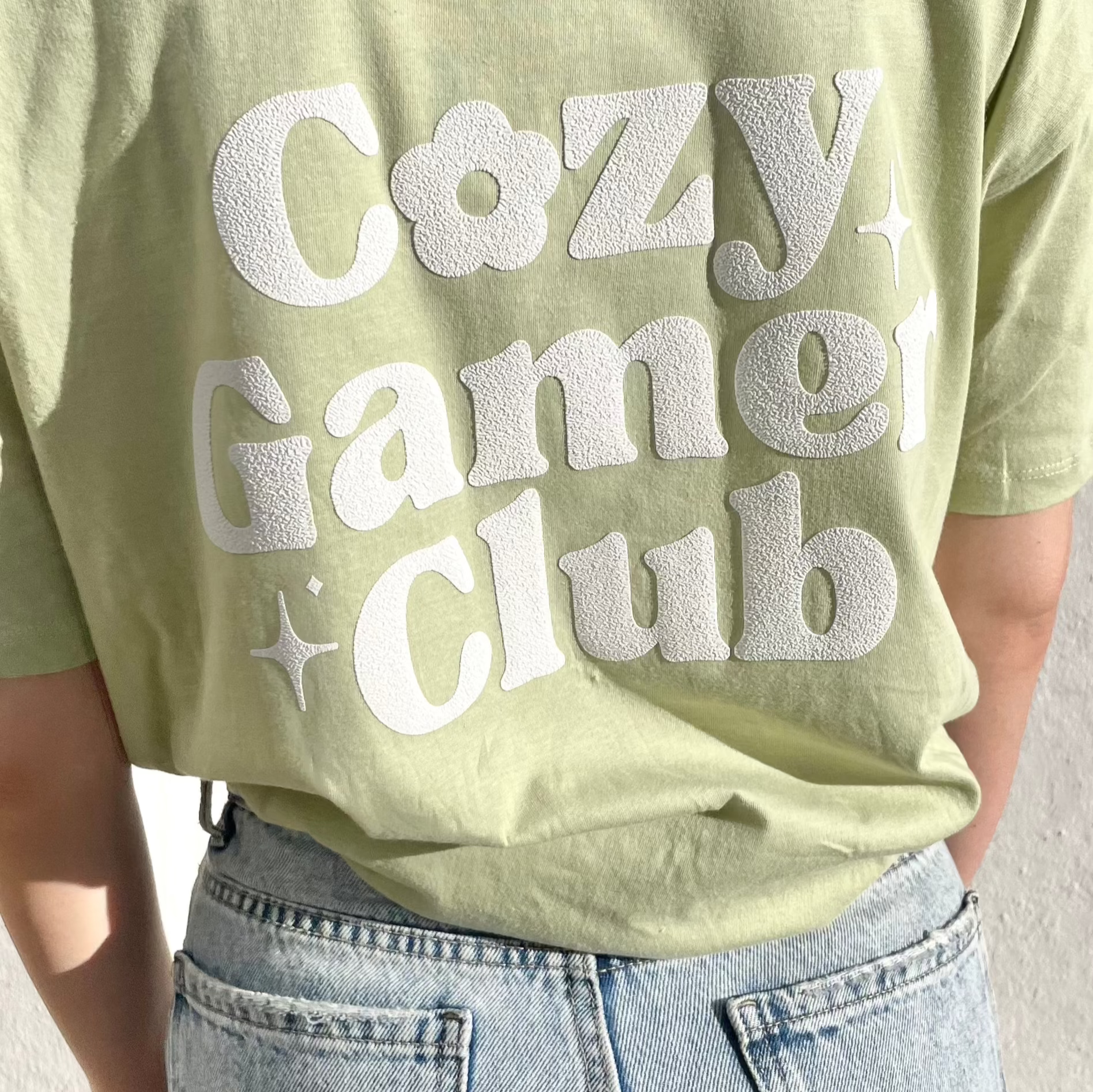 A close-up of a person donning a petimint Cozy Gamer Club T-Shirt in light green, featuring the text 