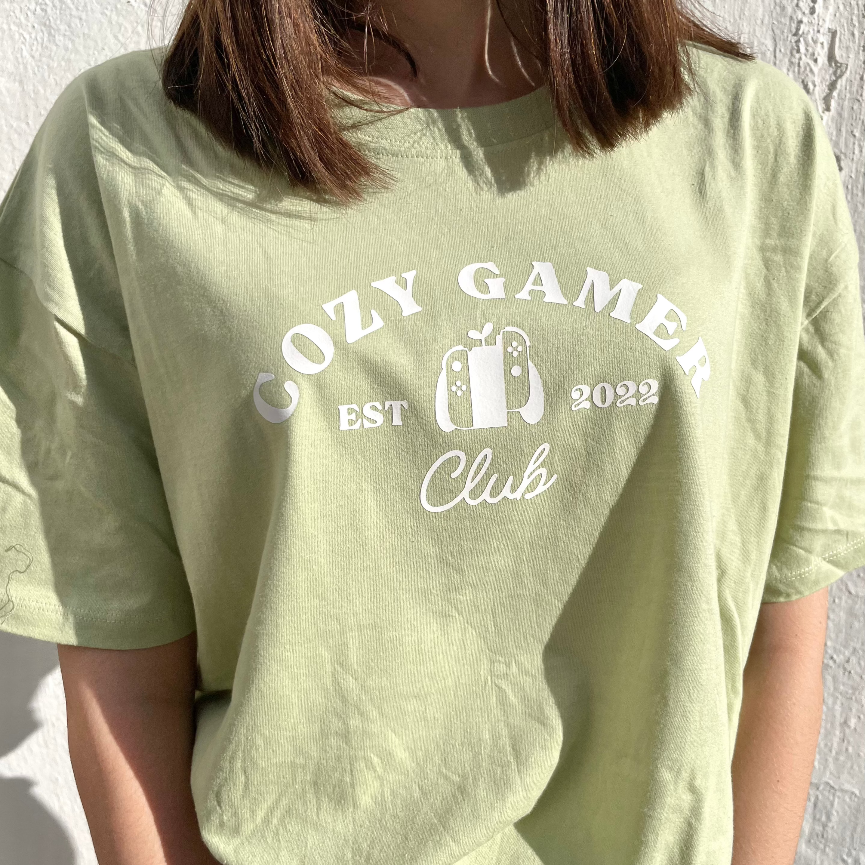 A person with shoulder-length brown hair wears the Pastel Green Cozy Gamer Club T-Shirt by petimint, featuring the text 