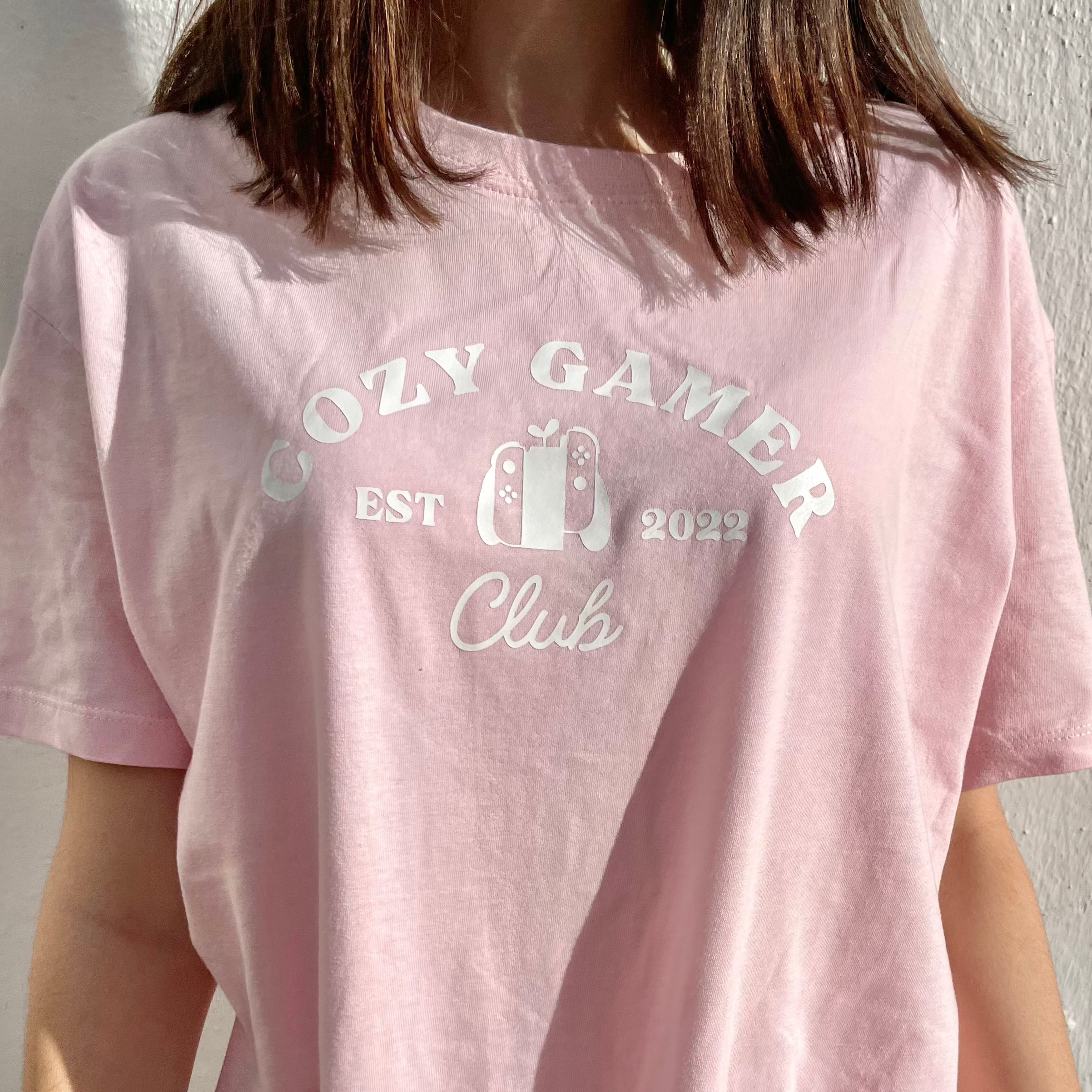 A person wearing a pastel pink Cozy Gamer Club T-Shirt by petimint, featuring the text 