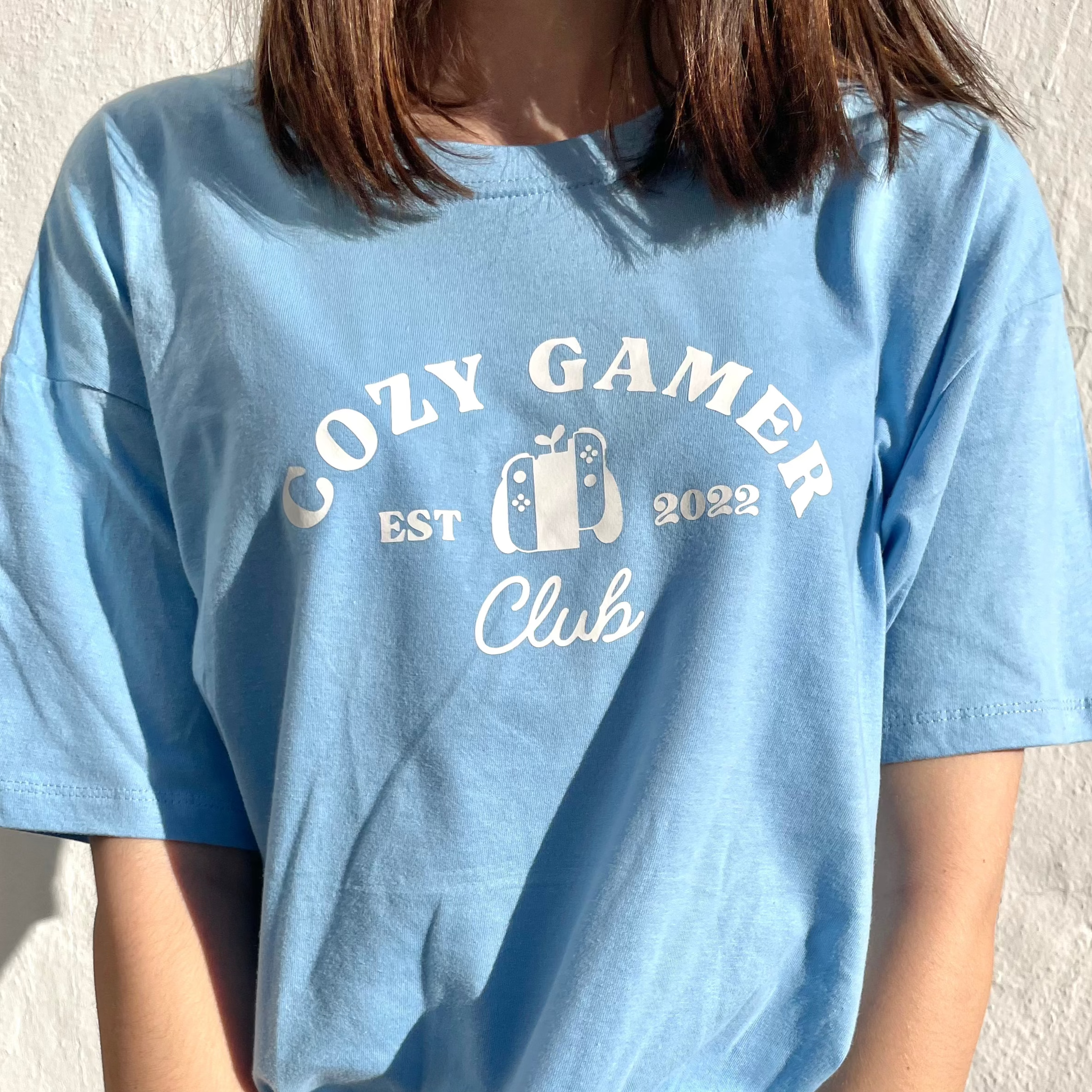 A person wearing a petimint's Pastel Blue Cozy Gamer Club T-Shirt, featuring the text 