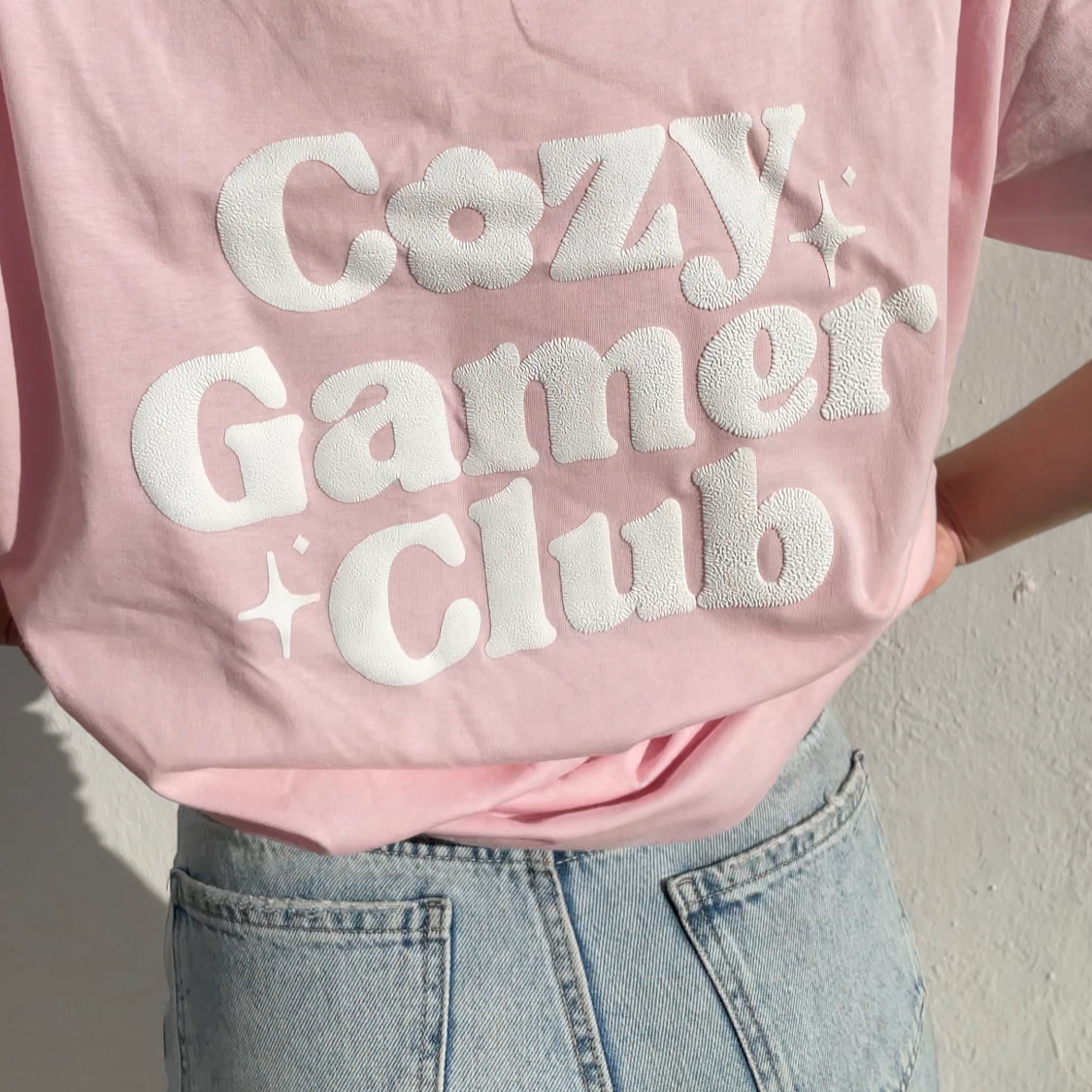 A person is wearing a petimint Cozy Gamer Club T-Shirt, which features the text 