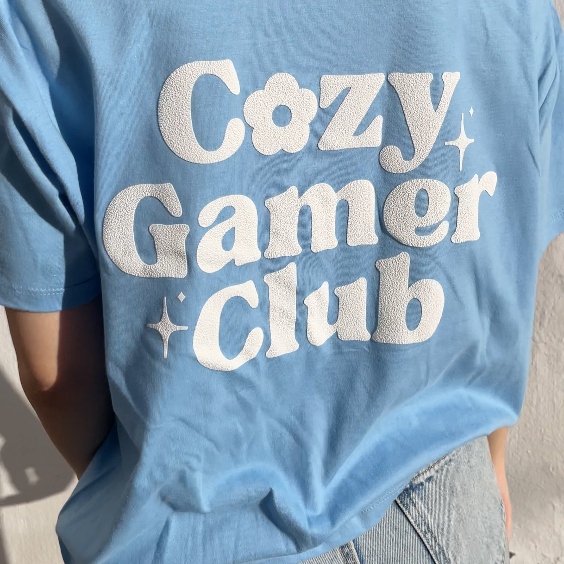 A person stands against a plain wall, evoking the relaxed charm of Stardew Valley, wearing petimint's Cozy Gamer Club T-Shirt in light blue, adorned with 