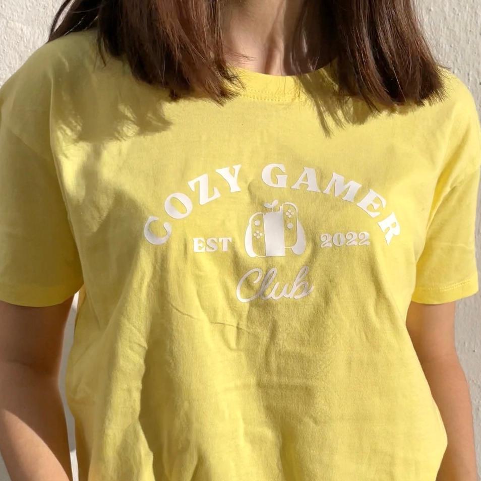 A person with shoulder-length brown hair wears a pastel yellow Cozy Gamer Club T-Shirt by petimint, featuring the text 