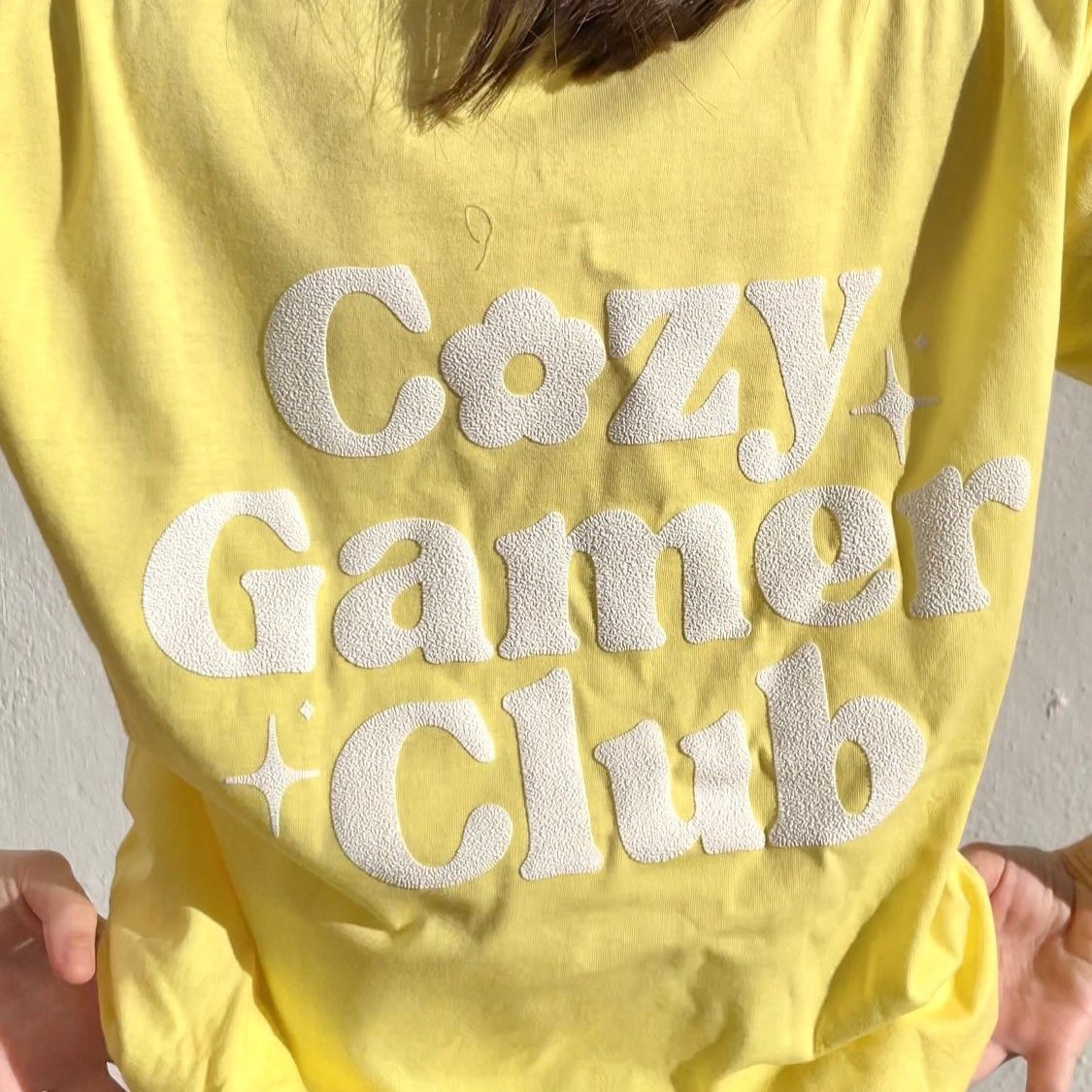 A person wearing the yellow Cozy Gamer Club T-Shirt by petimint, featuring textured white text that reads 