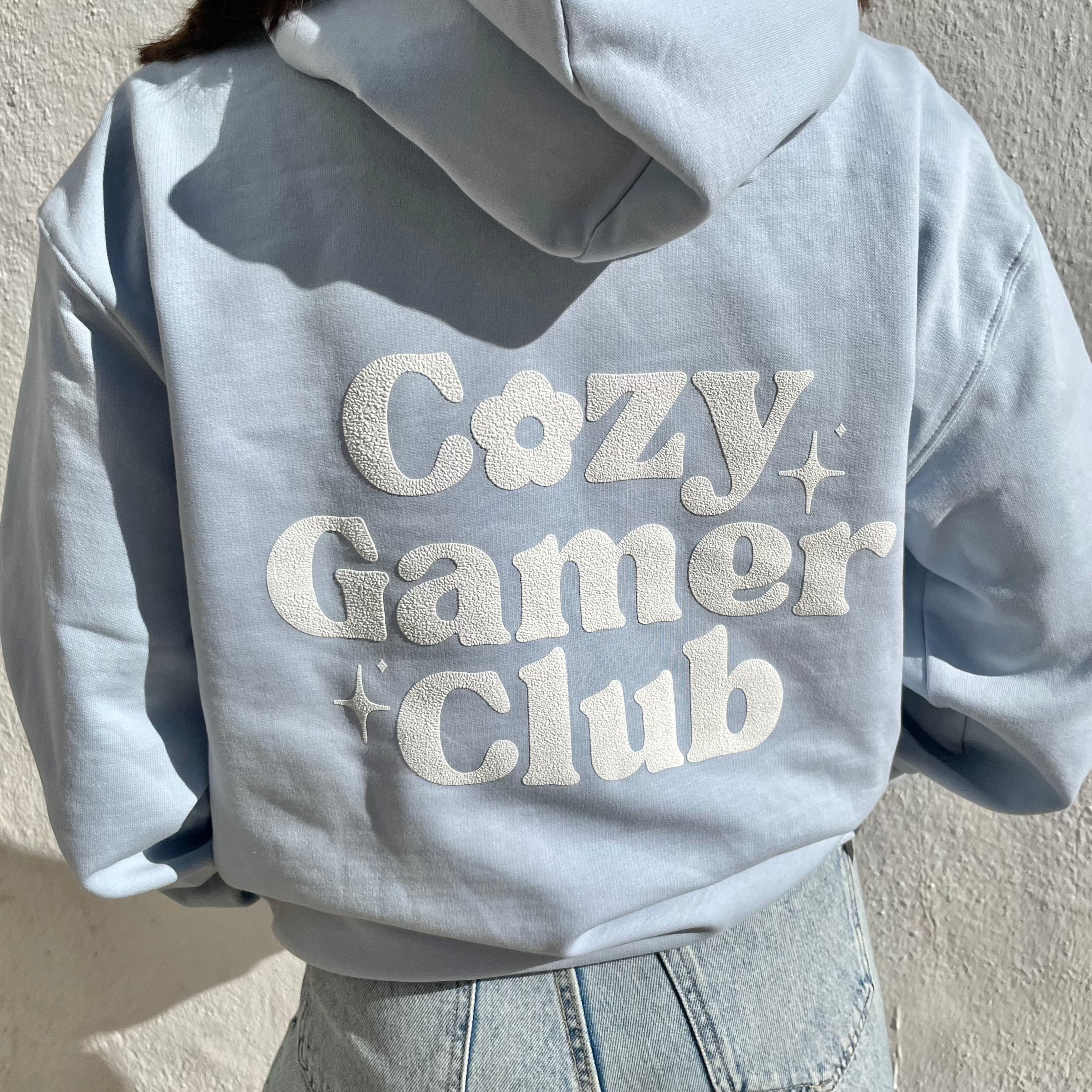 A person wearing a pastel blue 100% cotton "Cozy Gamer Club Hoodie" by petimint, featuring white text with playful flower and star designs on the back. This original design pairs perfectly with their light-washed jeans as they stand against a sunlit white wall.