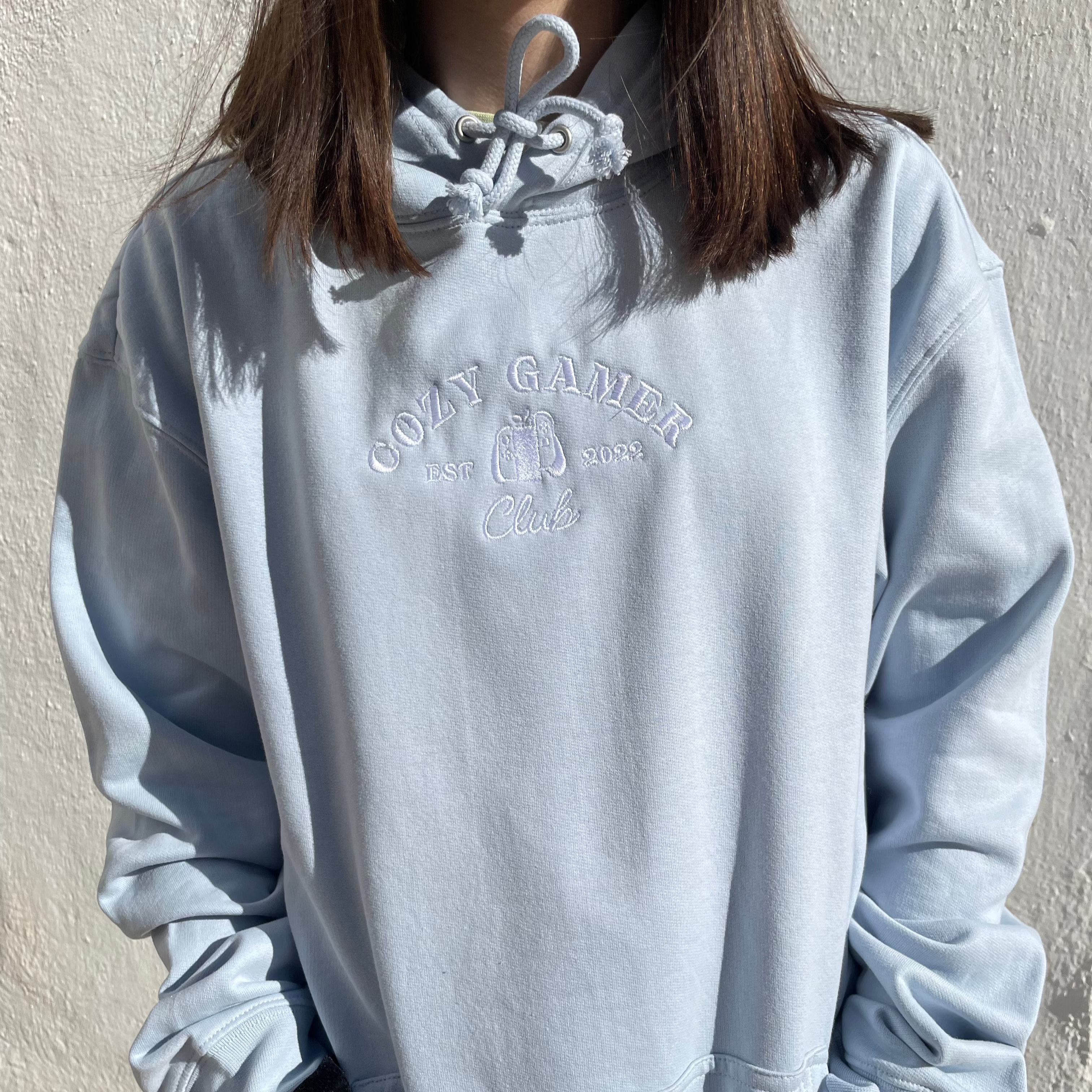 A person wearing a light blue petimint Cozy Gamer Club Hoodie, made from 100% cotton with "COZY GAMER Club EST. 2023" embroidered on the chest, stands against a white wall. The original design highlights the person's hands in their pockets, with their head cropped out of the image.