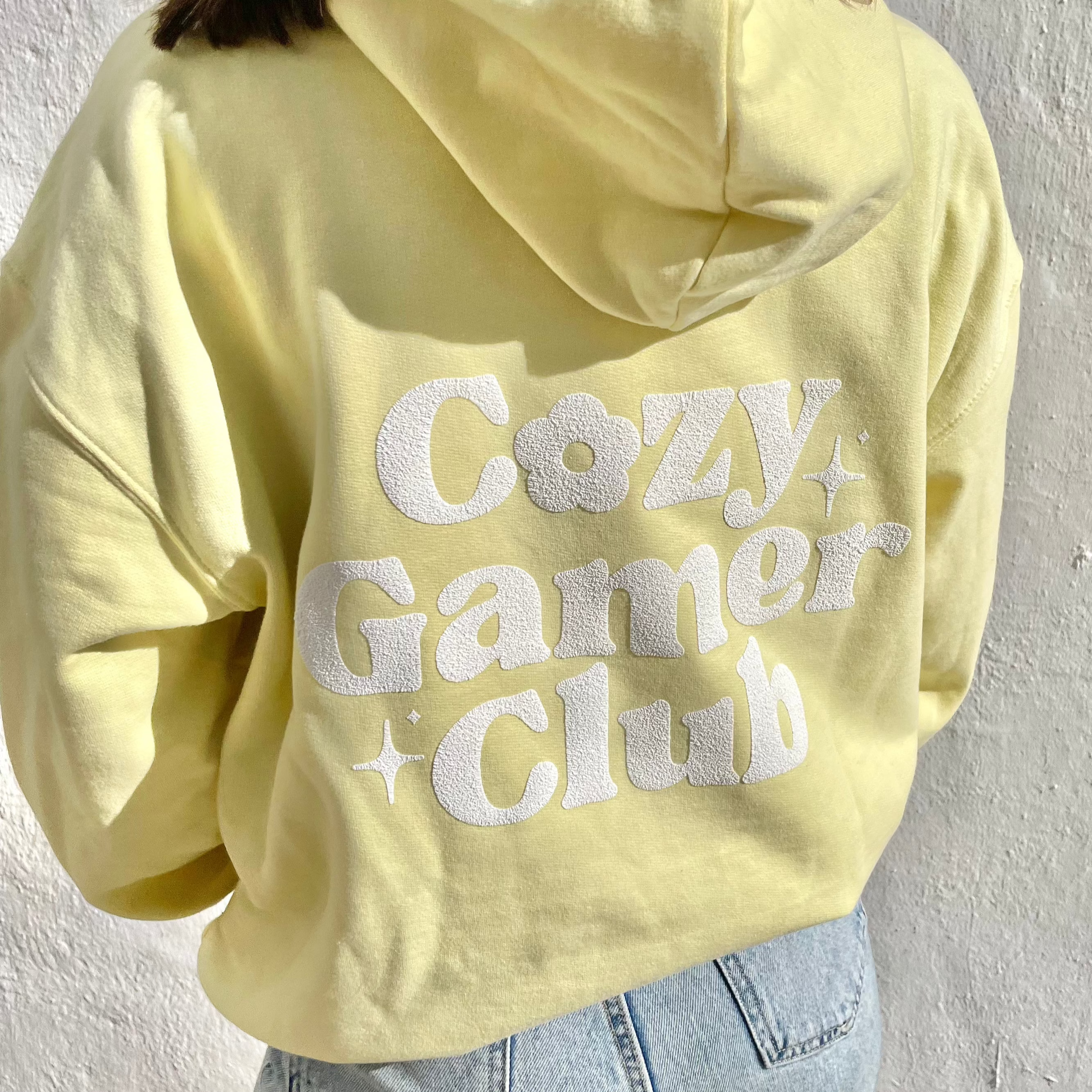 A person wearing a pastel yellow Cozy Gamer Club Hoodie by petimint, featuring white textured 