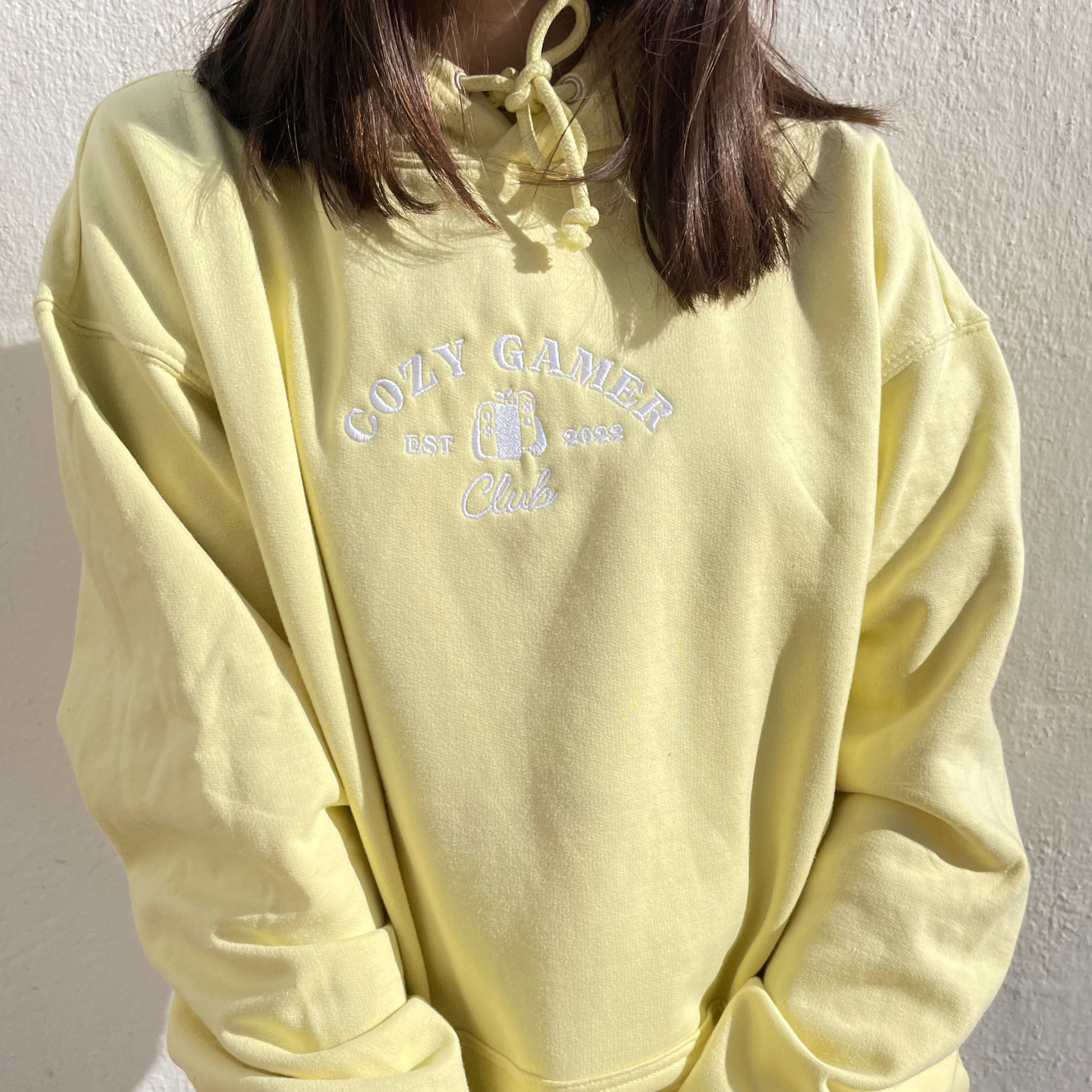 A person wearing a petimint Cozy Gamer Club Hoodie in light yellow, crafted from 100% cotton with white embroidery that reads 