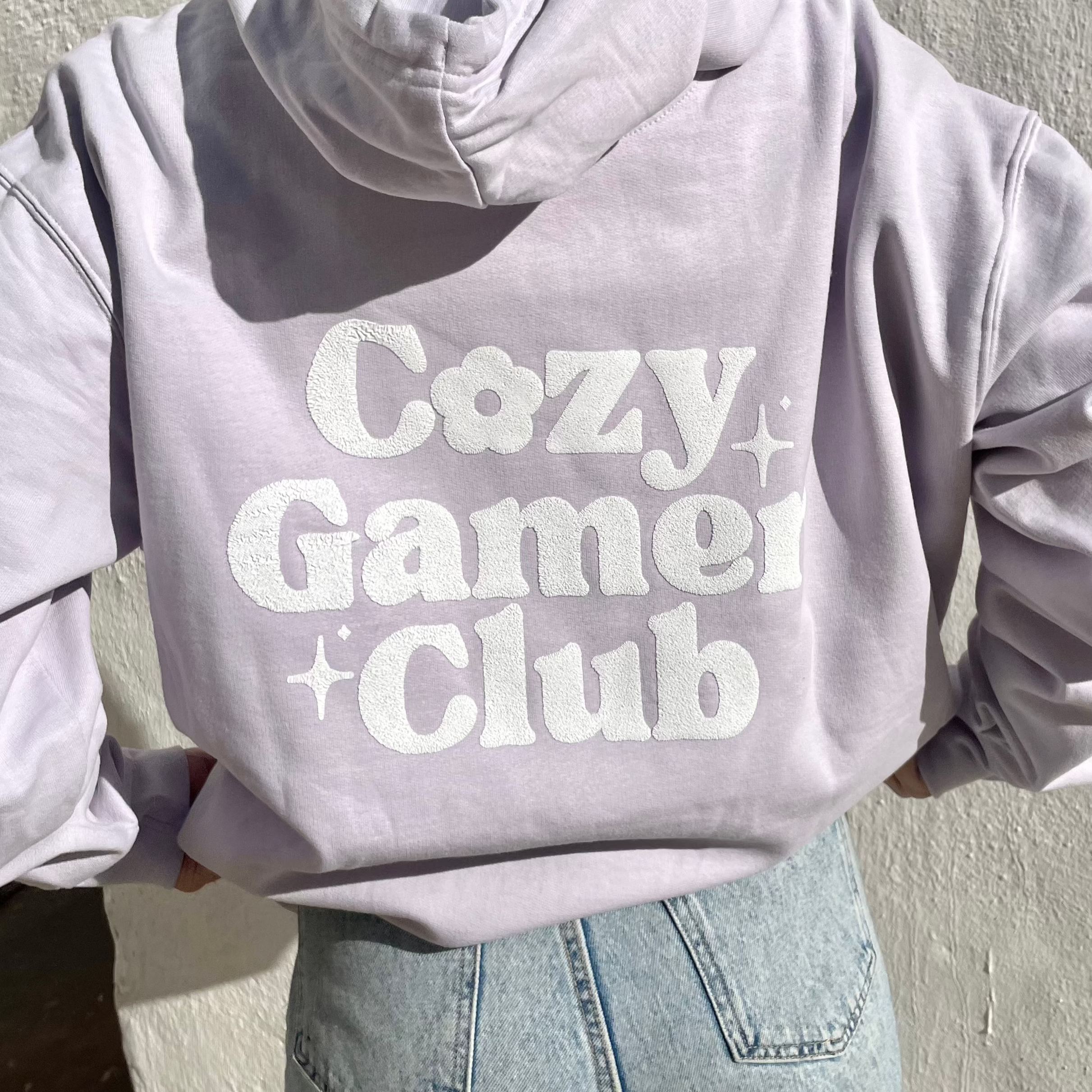 A person wearing a pastel lilac hoodie from petimint made of 100% cotton with the back design reading "Cozy Gamer Club" in large, white, playful letters. The Cozy Gamer Club Hoodie pairs perfectly with their light blue jeans as they stand against a white background.