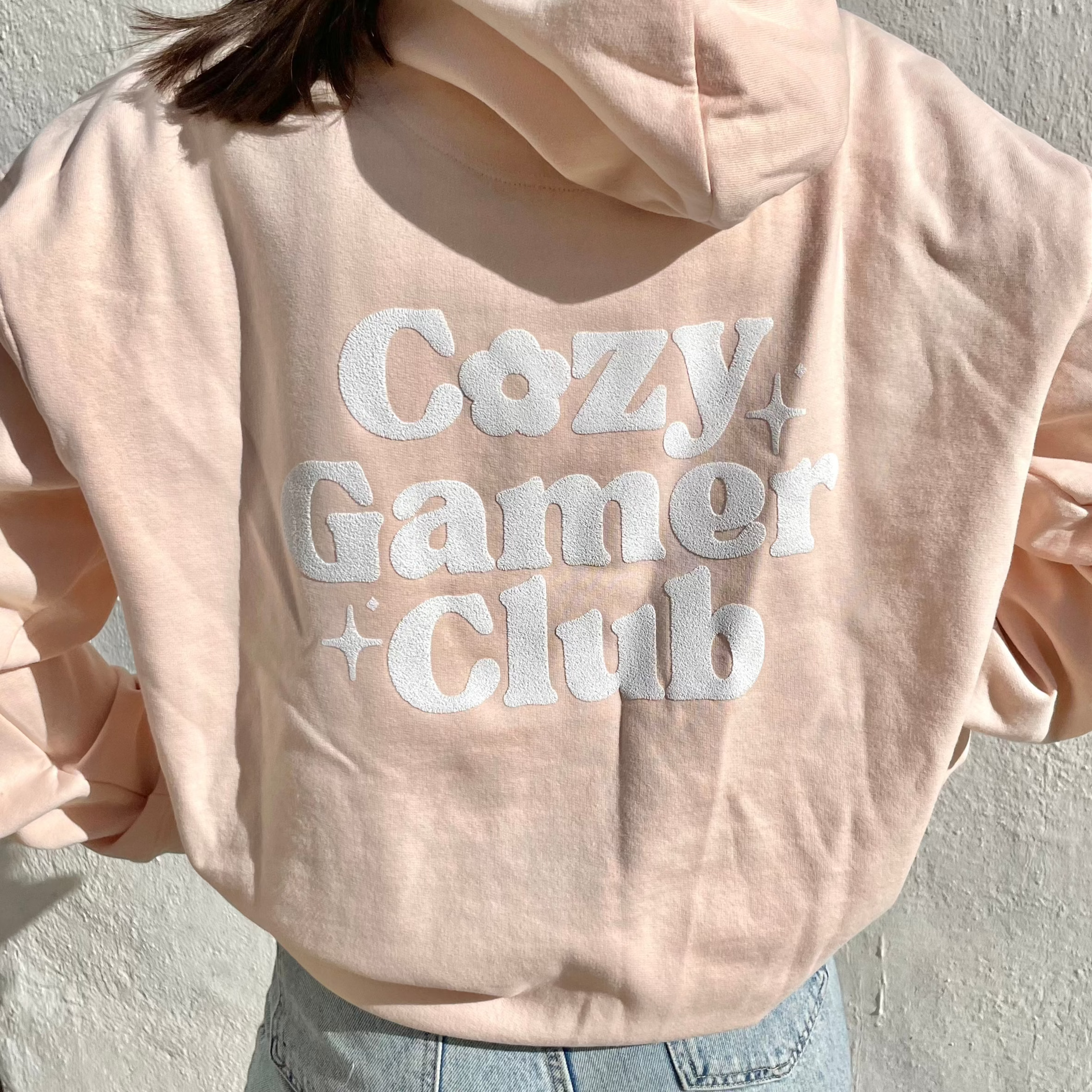 A person is wearing the Pastel Cream "Cozy Gamer Club Hoodie" by petimint, made from 100% cotton, featuring "Cozy Gamer Club" printed on the back in a white, textured, and playful font. As they pose against a plain white wall with only their back visible, their medium-length hair emphasizes the unique original design by Petimint.