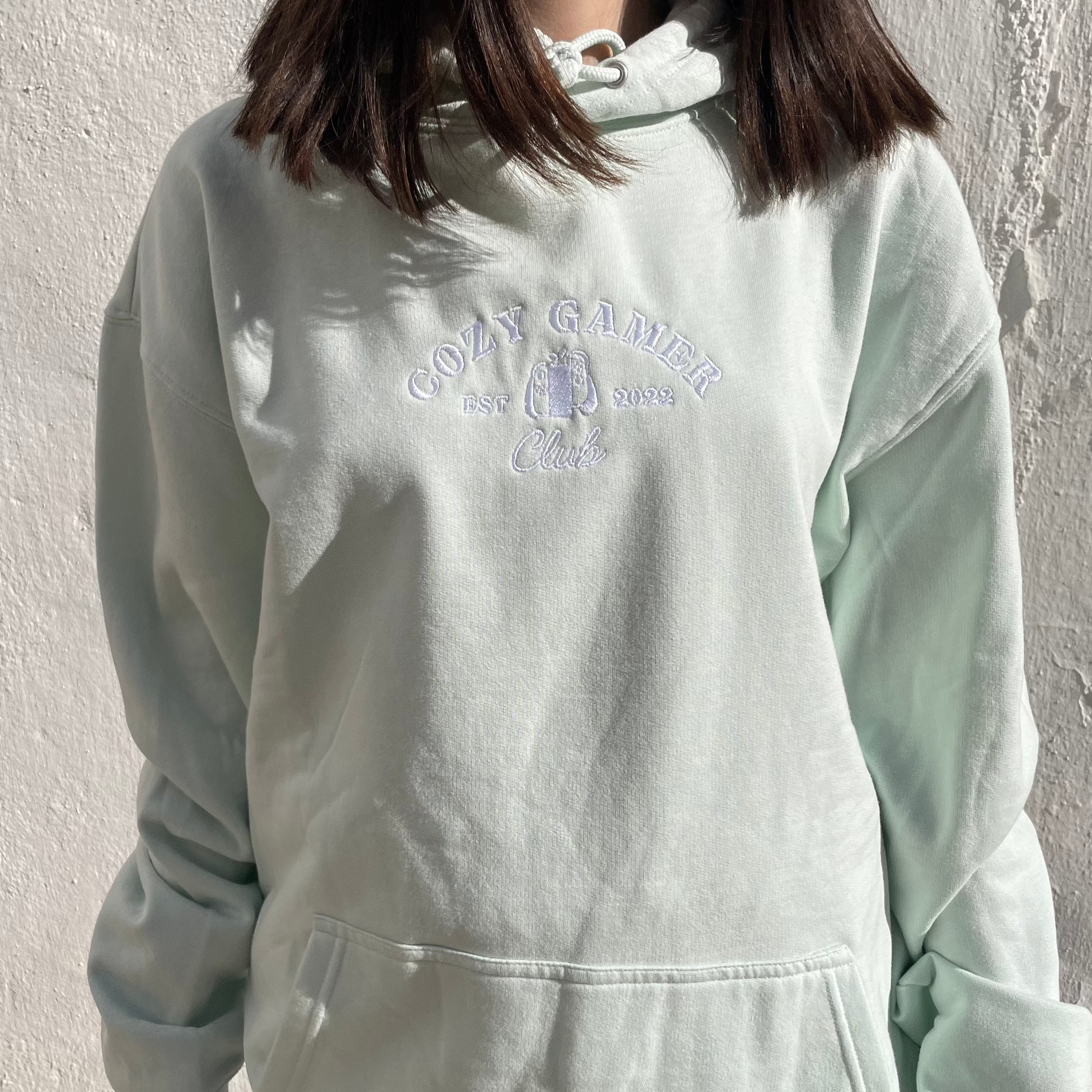 A person wearing a light green Cozy Gamer Club Hoodie by petimint, featuring embroidered text reading 