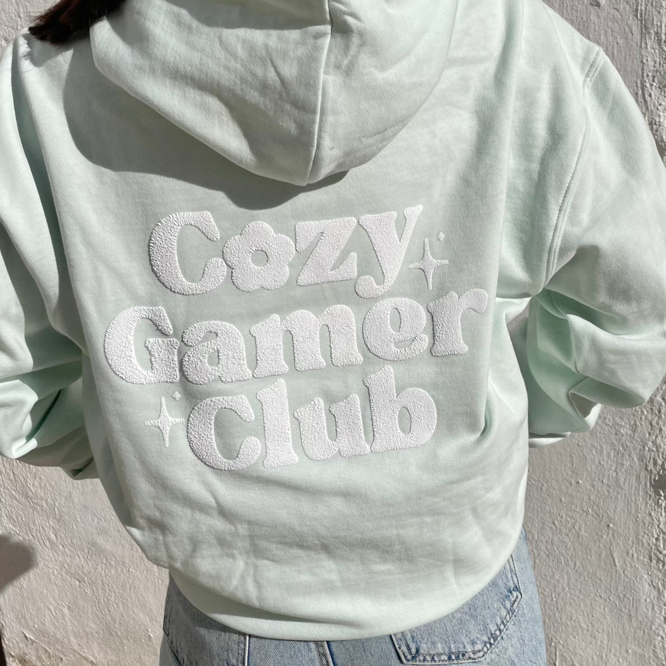 A person wearing a pastel mint Cozy Gamer Club Hoodie from petimint, adorned with the text 