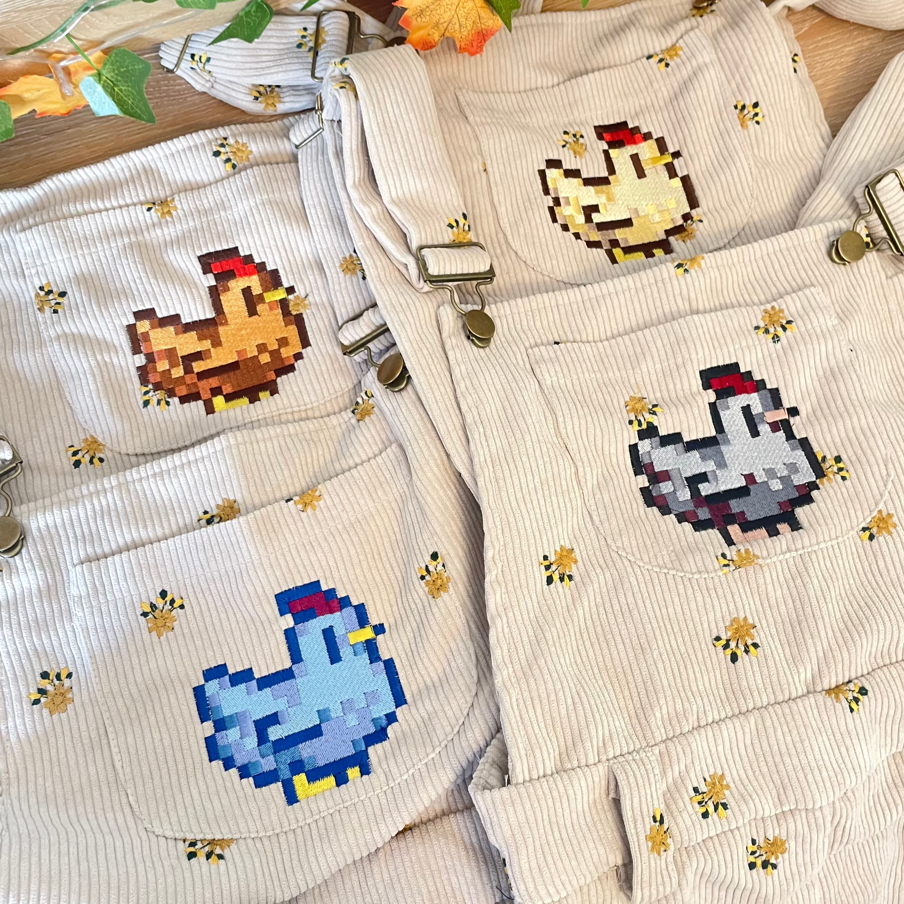 Stardew Chicken Floral Overalls Long Pants. A person stands against a beige wall, wearing a white T-shirt under the petimint Stardew Chicken Floral Overalls Long Pants. The light beige corduroy overalls feature a pixelated blue bird design on the front bib and small floral patterns spread throughout, giving off major cozygamer vibes. The person's head is not visible in the image.