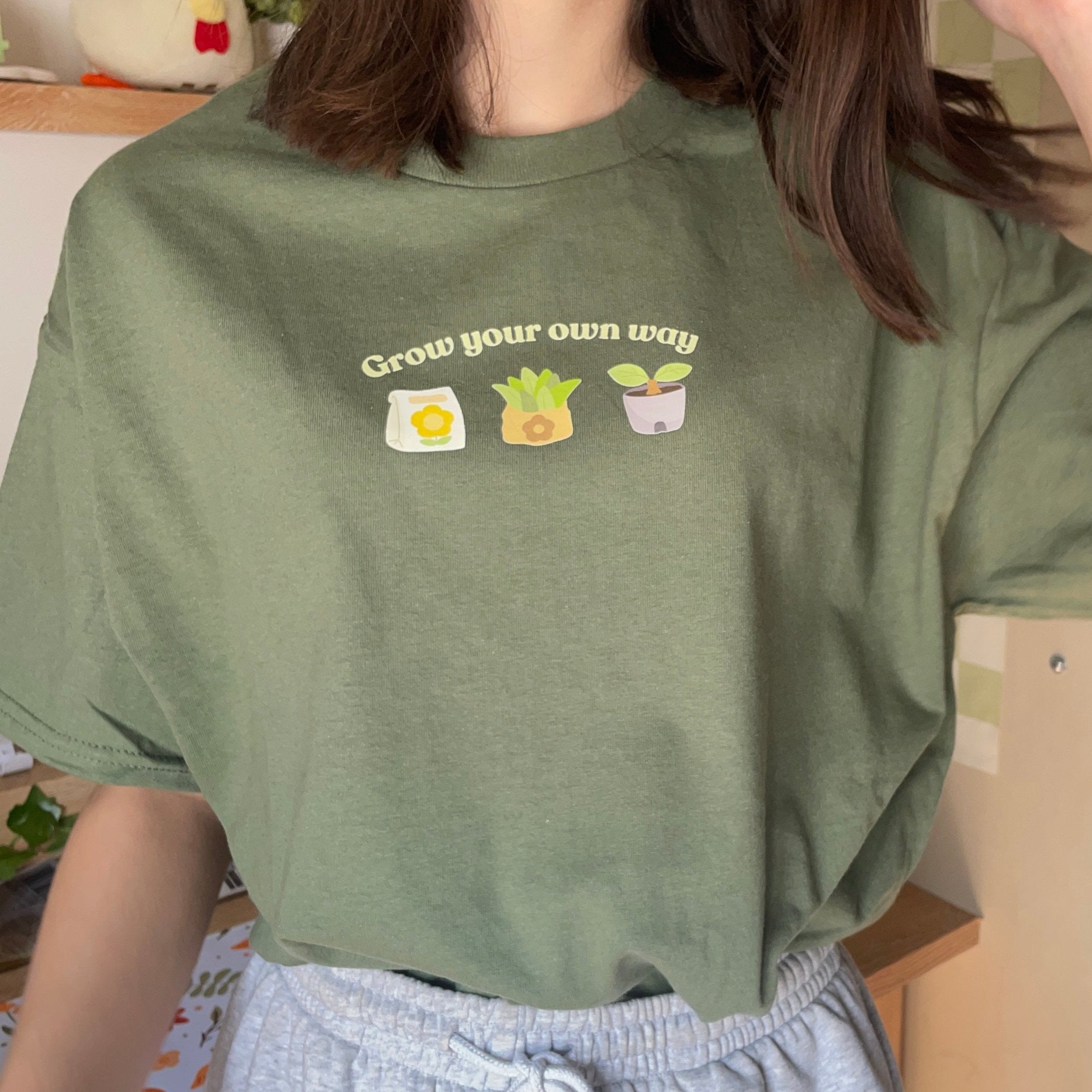 A person wearing a petimint's "Grow your own way ACNH T-Shirt" adorned with illustrations of a bag of soil, a potted plant sprout, and a potted plant, bringing to mind images from Stardew Valley. They are also dressed in light gray pants and are standing indoors amidst various objects in the background.