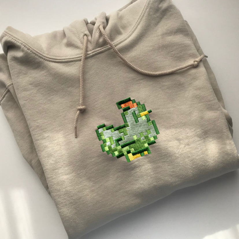 The Stardew Chicken Green Hoodie, by petimint, is neatly folded with a drawstring hood. On the front of the light gray hoodie, there is an embroidered pixel art design of a green duck with an orange beak and feet, making it perfect gaming apparel. The hoodie is displayed against a plain white background.