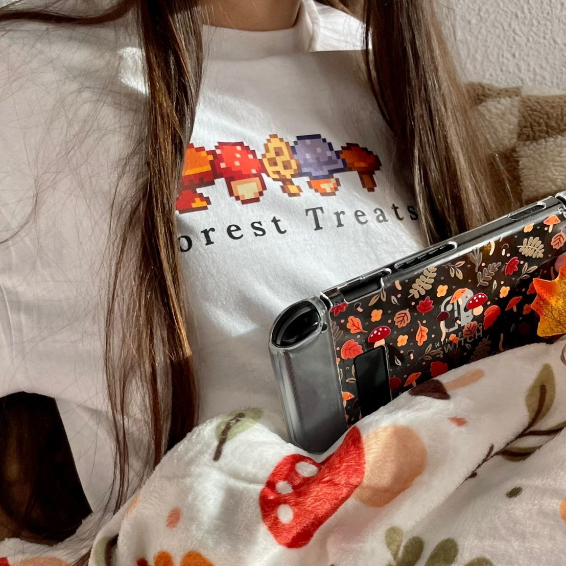 A person with long hair is wearing a white *petimint* Forest Treats T-shirt featuring pixel art of mushrooms and the text "Forest Treats." They are holding a handheld gaming console with a colorful mushroom-themed cover. This cozy gamer is also covered with a blanket adorned with mushroom patterns.