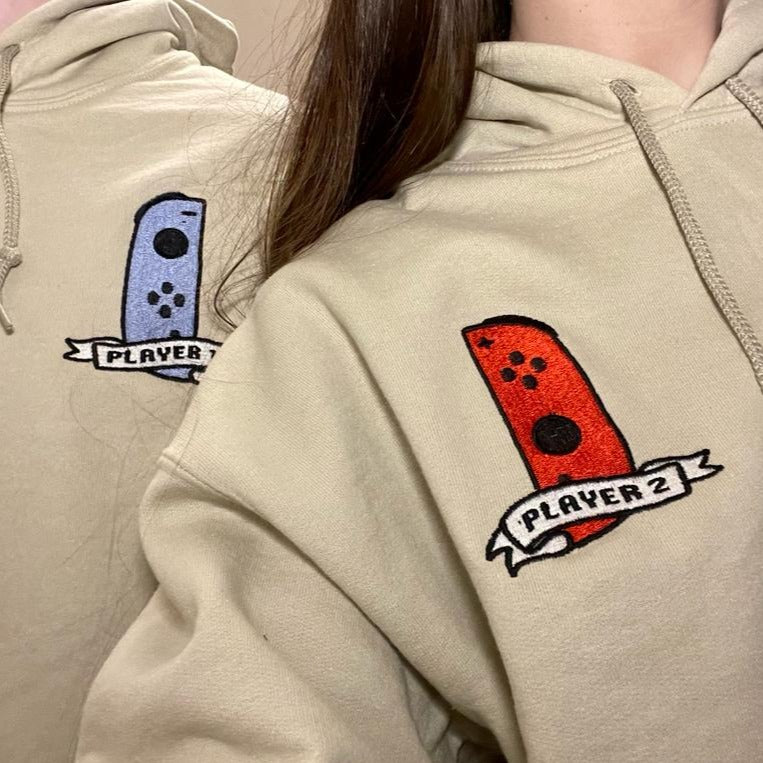 Two people are wearing matching hoodies from the petimint Pack 2 Hoodie Player 1 & 2 Gamer Apparel set. Each hoodie is embroidered with a design that features a game controller and a ribbon underneath. One ribbon reads 