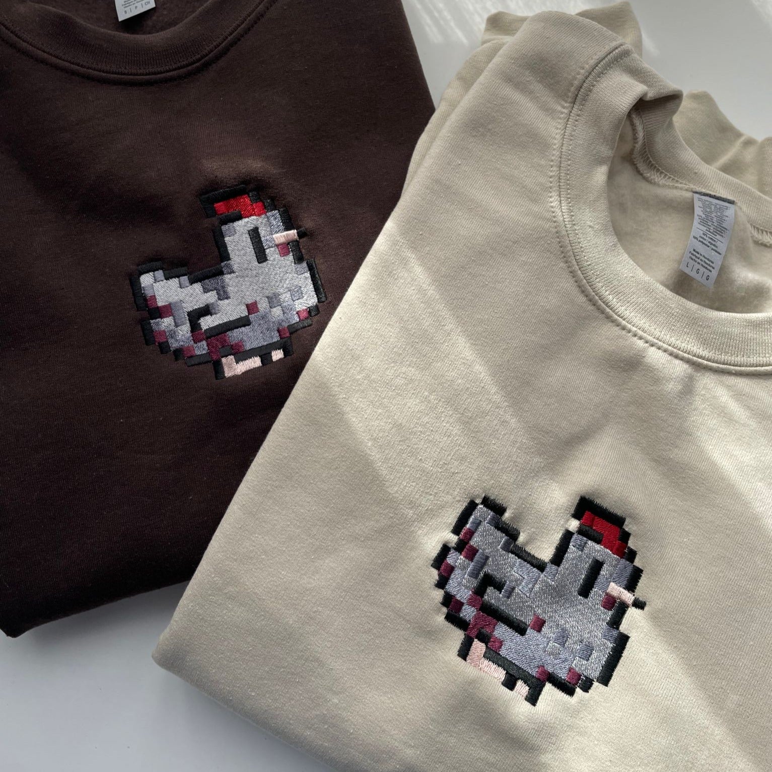 Two Stardew Black Chicken Crewneck sweatshirts by petimint, featuring pixel art-style chicken designs, are laid flat on a white surface. One sweatshirt is dark brown and the other is light beige. Both pieces of apparel showcase identical pixelated chickens with gray bodies, red combs, and black eyes positioned in the center.