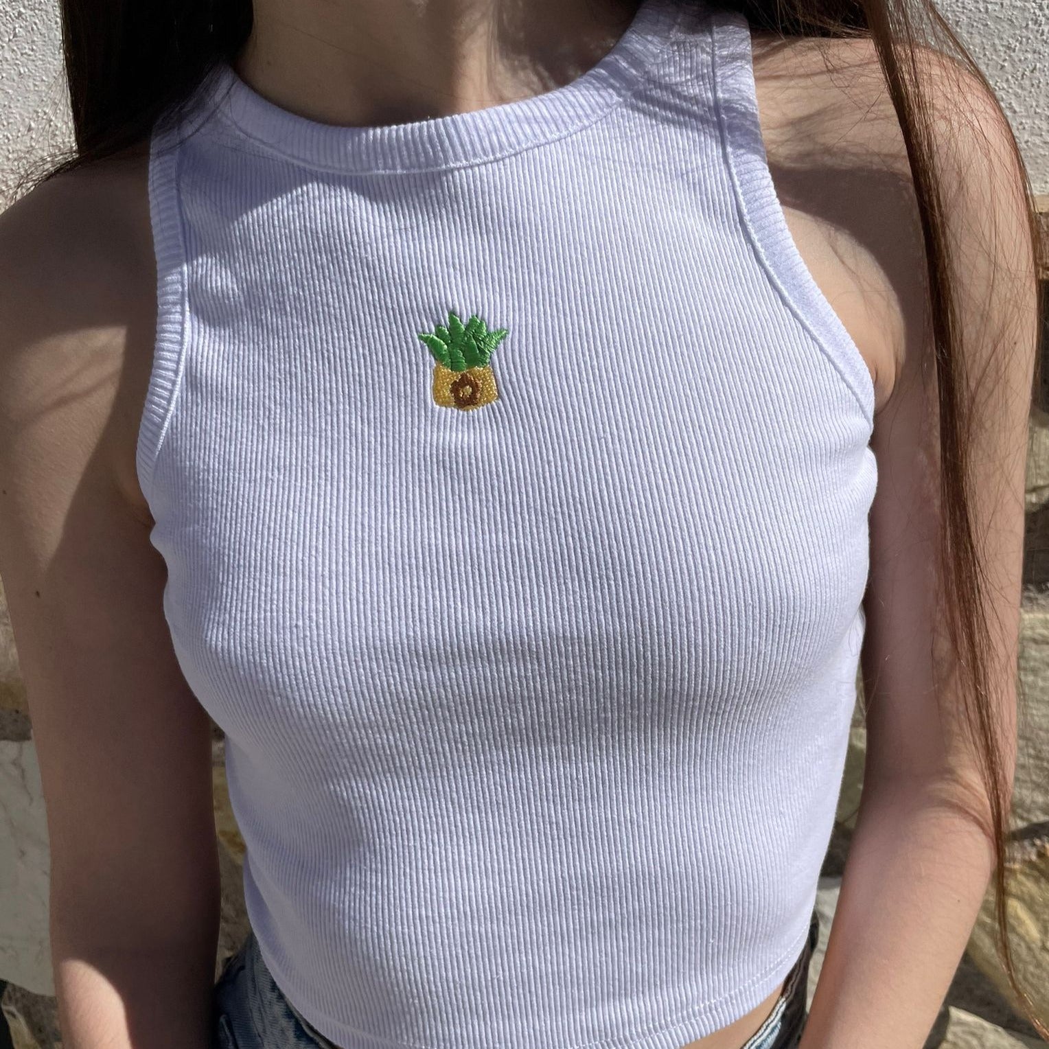 A person with long hair wears a sleeveless, white ribbed crop top from petimint's Cottage Tops Cozy Gamer Apparel collection, featuring a small embroidered pineapple with green leaves on the chest. The cozy gamer's background shows a stone wall in natural light, reminiscent of Stardew Valley's charming countryside.