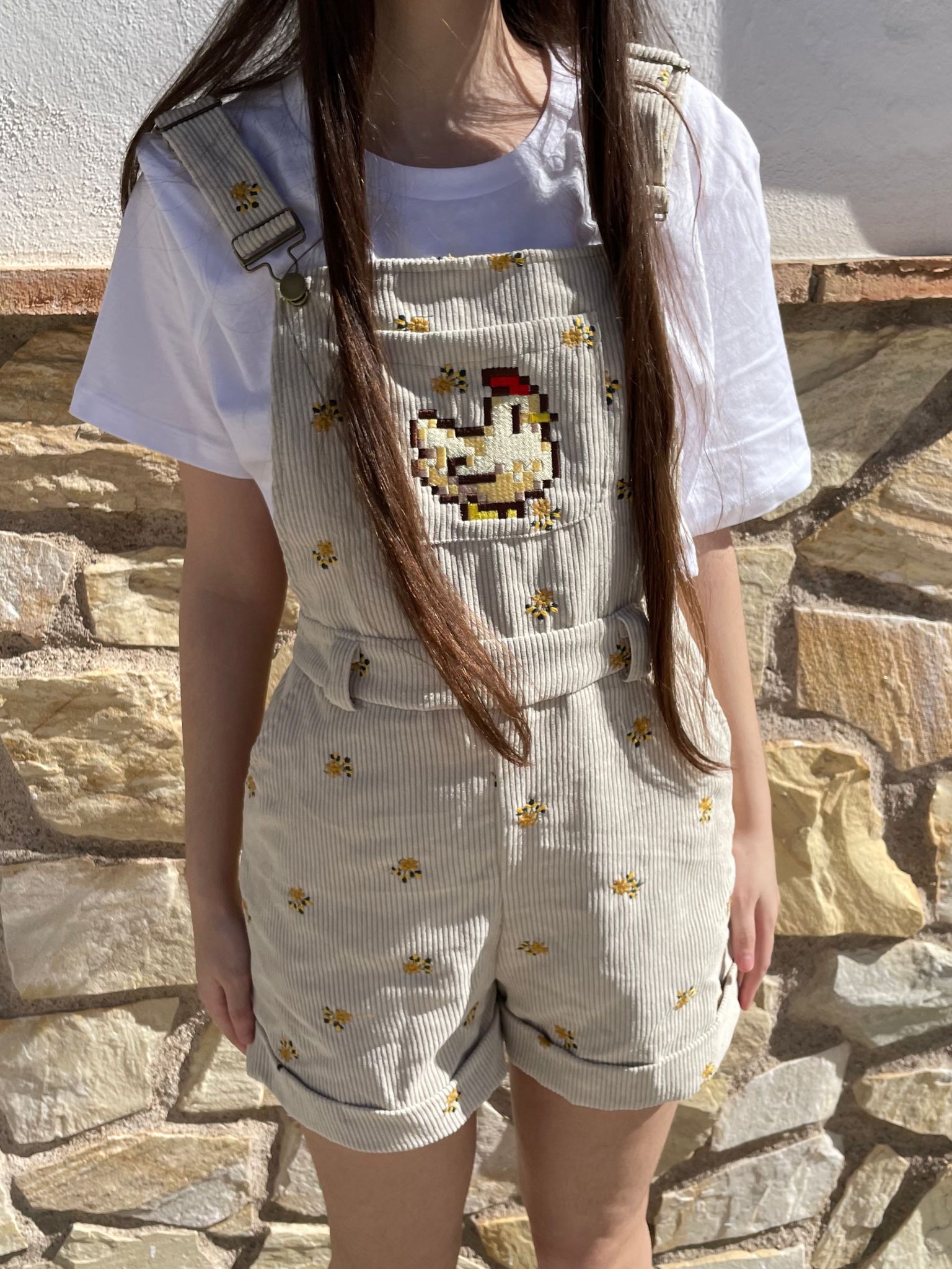 A person with long brown hair is wearing a white T-shirt paired with the Stardew Chicken Floral Overalls Shorts by petimint, which feature light gray fabric adorned with small yellow flower patterns and a pixelated chicken design on the chest, adding a touch of cozygamer charm. The background shows a stone wall and part of a white building. The person's face is not visible.