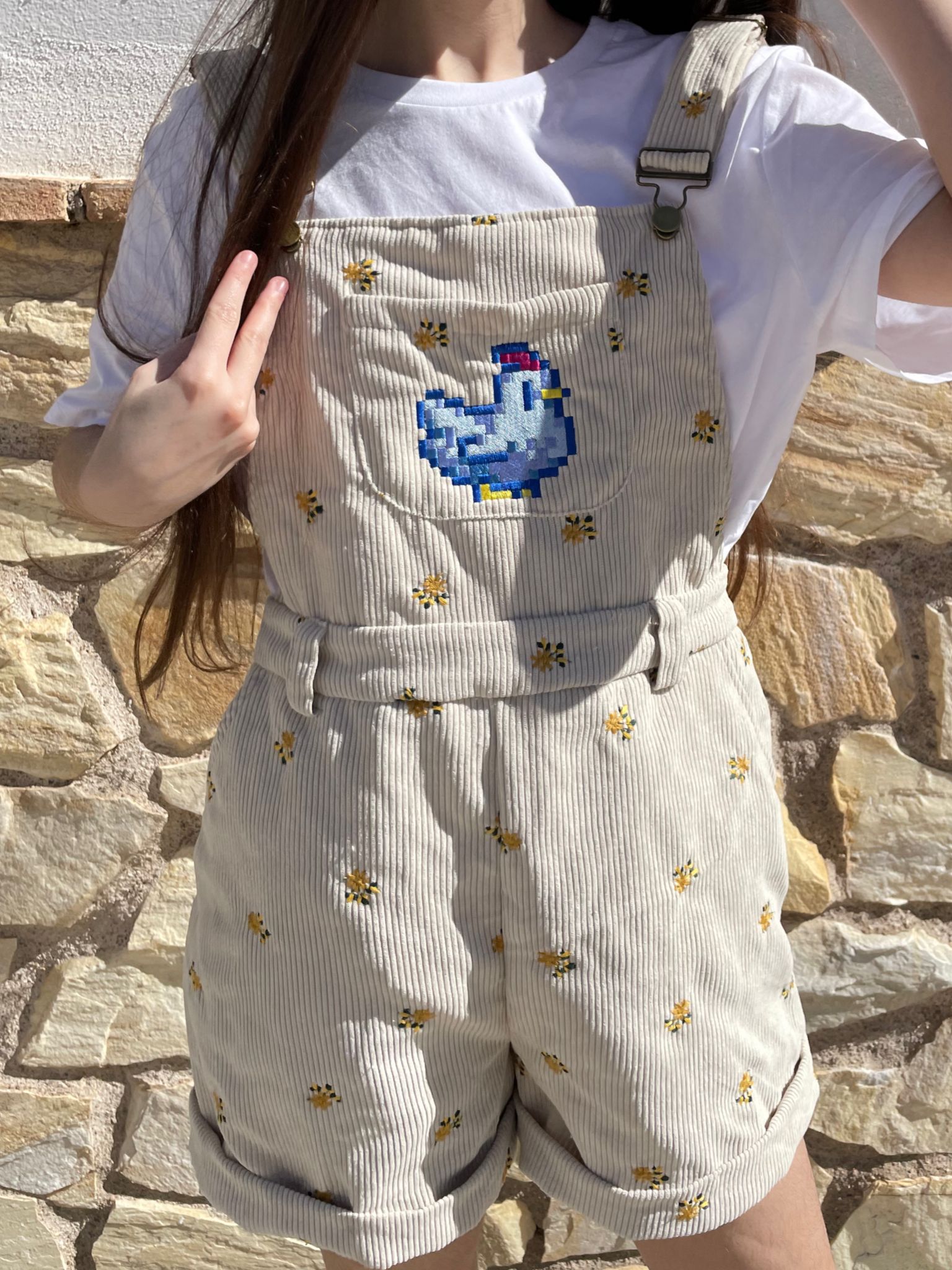 A person with long hair is wearing a white t-shirt under petimint's Stardew Chicken Floral Overalls Shorts, adorned with a blue duck and flower embroidery design. Standing against a stone wall and making a peace sign, they exude cozygamer vibes, seemingly ready for some quality time with their Nintendo Switch.