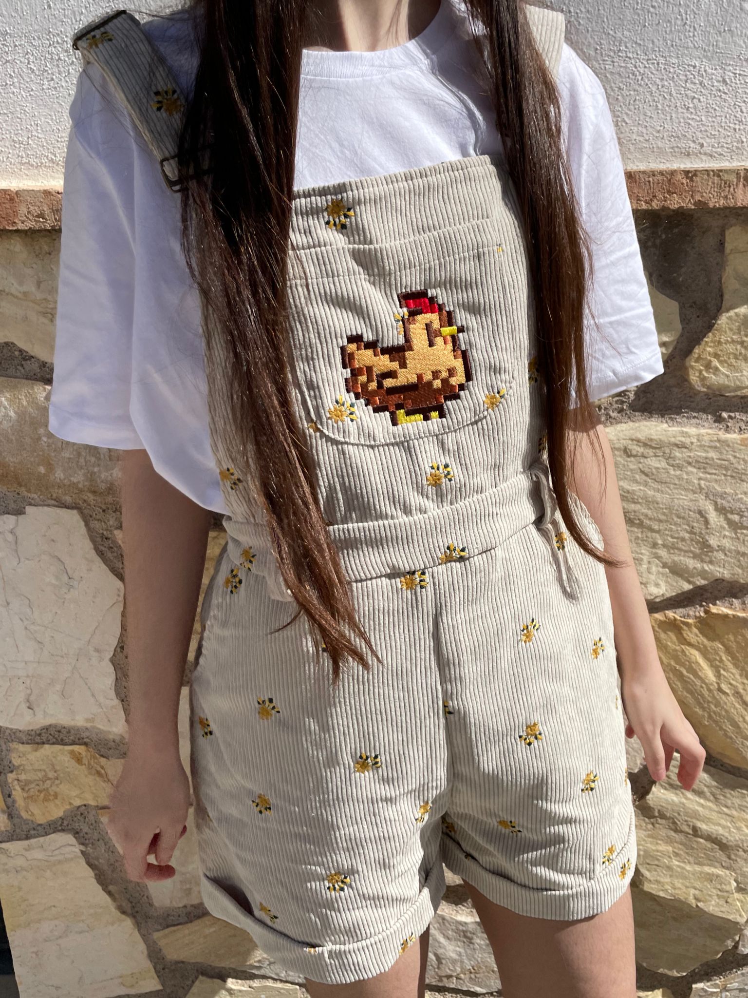 A person wearing a petimint white t-shirt and the Stardew Chicken Floral Overalls Shorts, embellished with small yellow chick patterns, stands against a stone wall while holding a Nintendo Switch. The overalls have a large embroidered chicken on the chest, and their long brown hair flows freely.