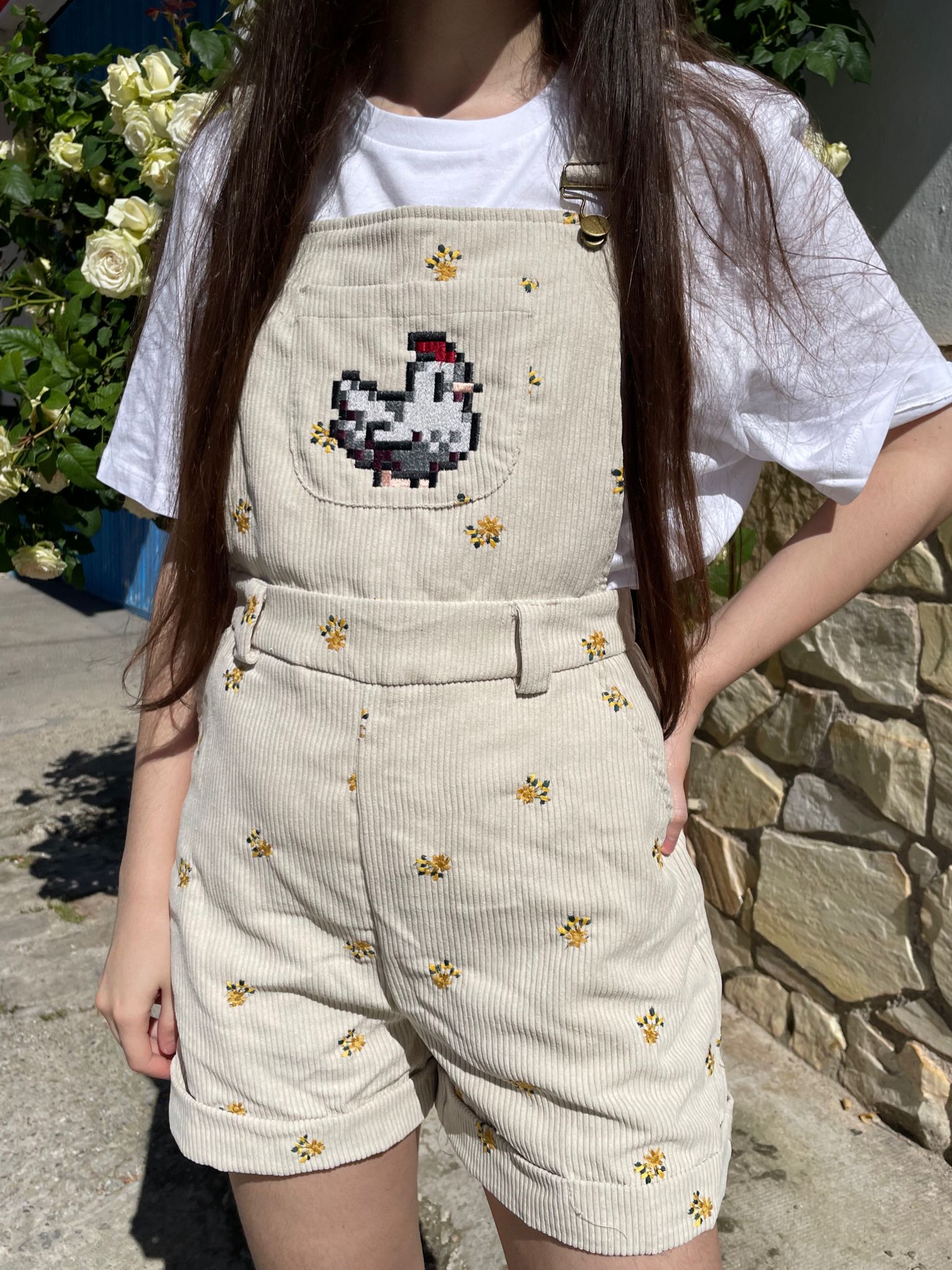 A person is wearing the Stardew Chicken Floral Overalls Shorts from petimint, featuring cream-colored corduroy with embroidered yellow flowers and a pixelated chicken reminiscent of gaming art on the front pocket. They are standing outdoors in front of green foliage and a stone wall, paired with a white t-shirt underneath the overalls.