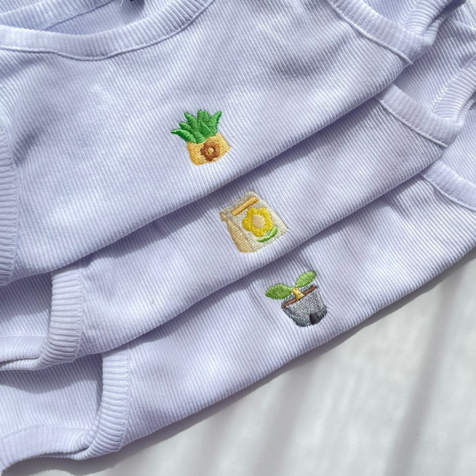 Three white ribbed shirts from petimint’s Cottage Tops Cozy Gamer Apparel collection are stacked on top of each other, each showcasing a small embroidered design: a pineapple, a jar with a lemon, and a plant in a pot. These charming pieces are displayed in a gently lit environment, evoking the cozy ambiance found in Stardew Valley.