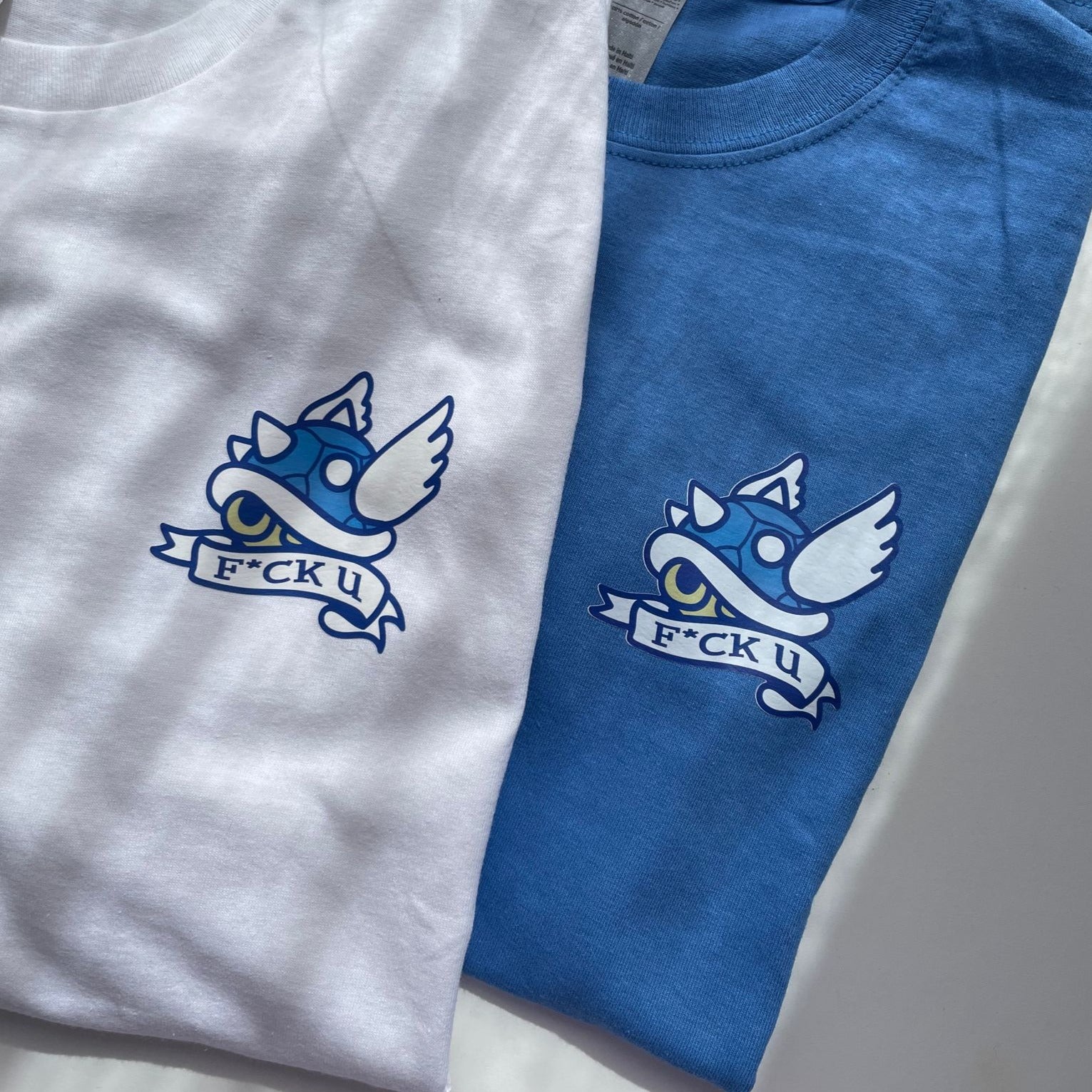 Two cozy petimint F*ck u T-Shirts lie flat, one white and one blue. Each shirt features a blue spiked shell with wings and a ribbon below it displaying the text 