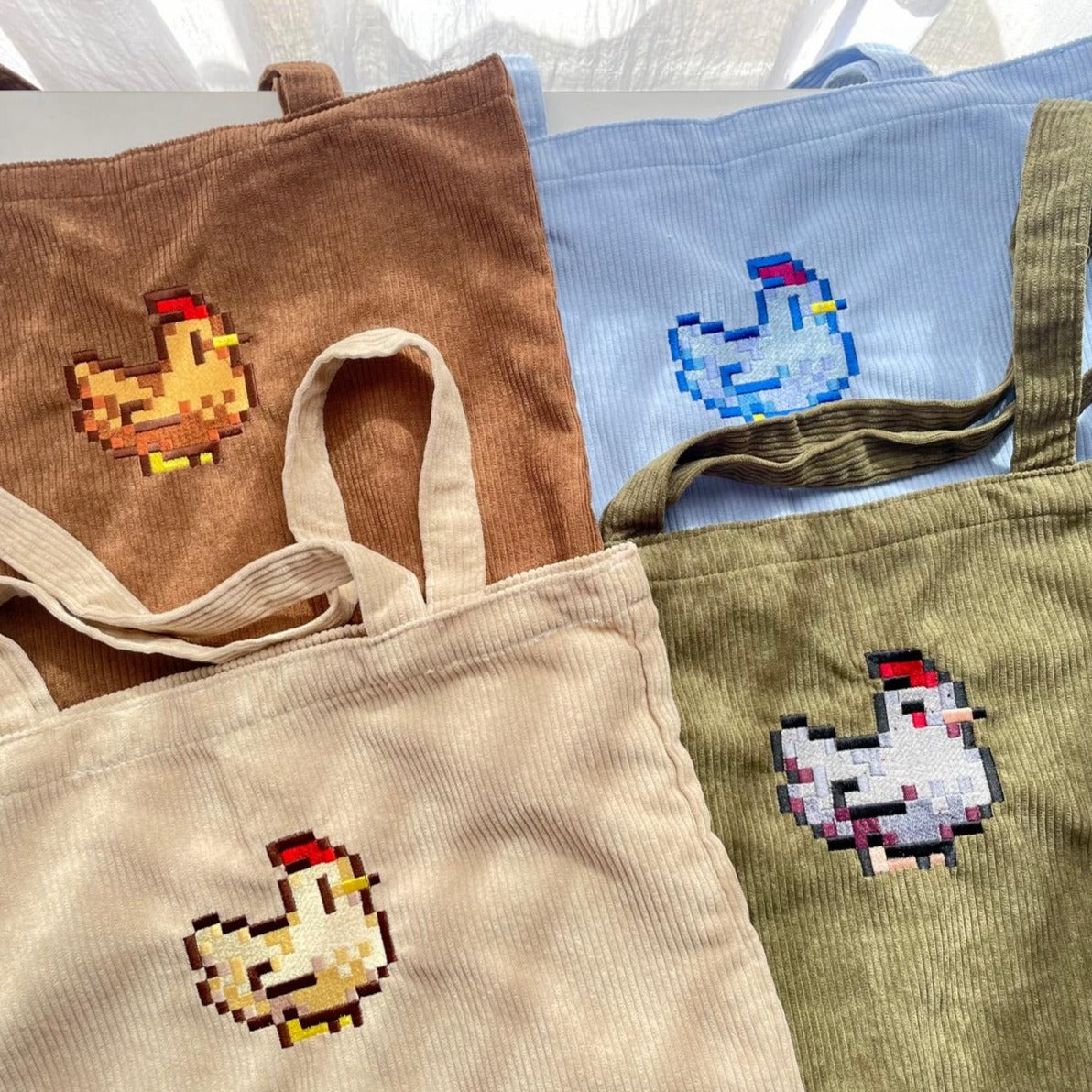 The petimint Stardew Chicken Tote Bags, with their corduroy texture and pixelated chicken designs, come in four colors: brown, blue, beige, and olive green. Each bag features a distinct chicken graphic adorned with embroidery. Sunlight creates gentle shadows on the fabric.