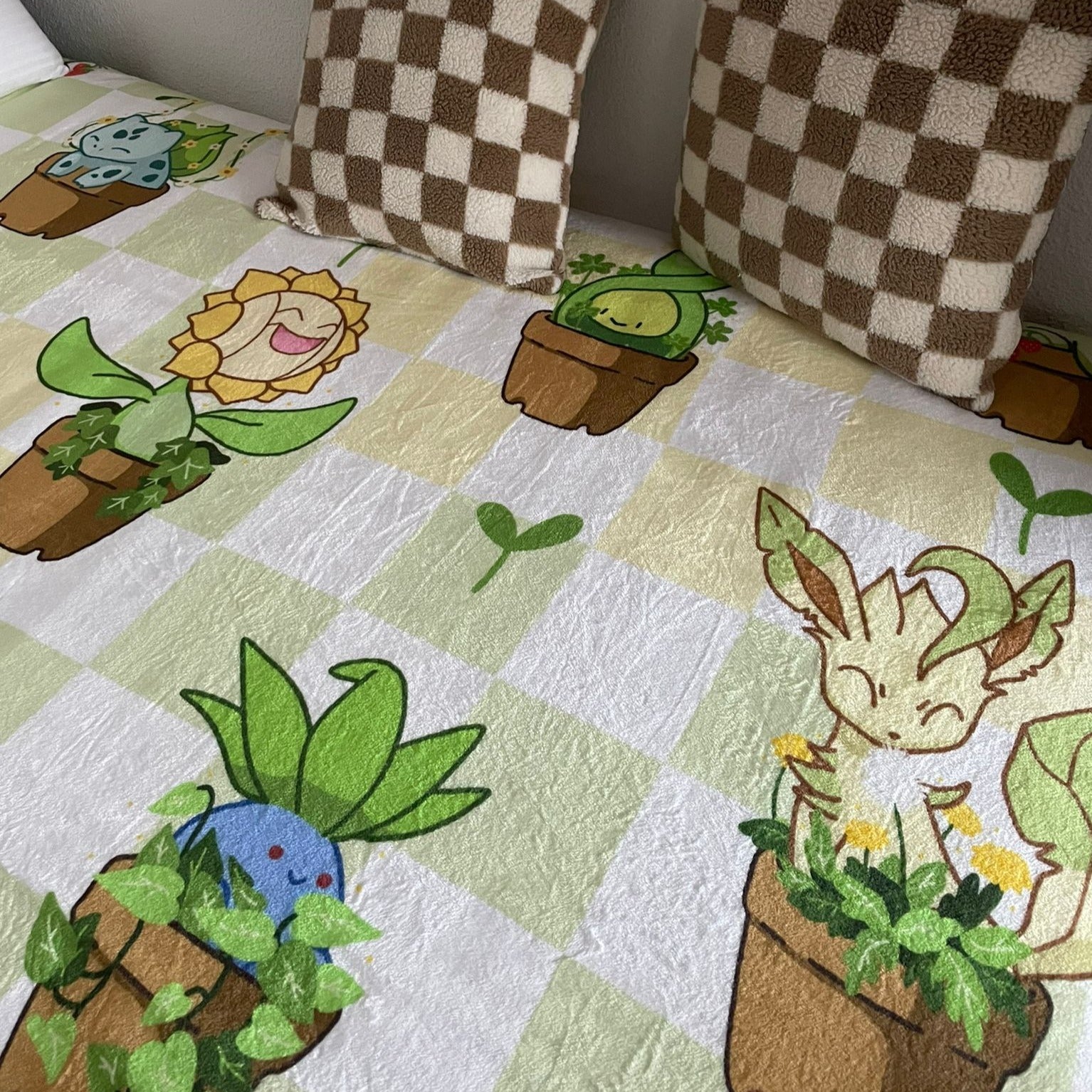 A bed is neatly made with the Pokepots Blanket Soft and Cozy by petimint, featuring a vibrant design of potted plants and charming characters with plant motifs, perfect for any cozygamer. Two checkered pillows at the head of the bed beautifully complement the checkered pattern on the blanket.