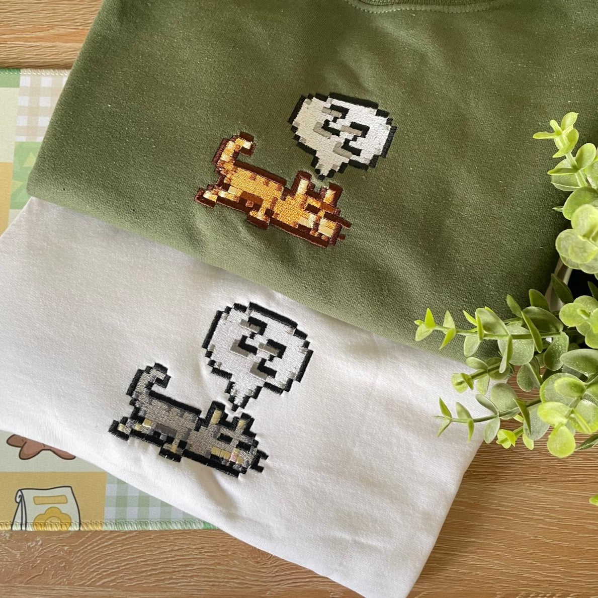 Two folded items of petimint's *Sleeping Cat Crewneck Apparel* are layered on a wooden surface, one green and one white, each showcasing a pixel art design of a cat with a speech bubble. A small green plant is positioned to the right, adding a touch of nature to the scene—a perfect setup for any cozygamer.