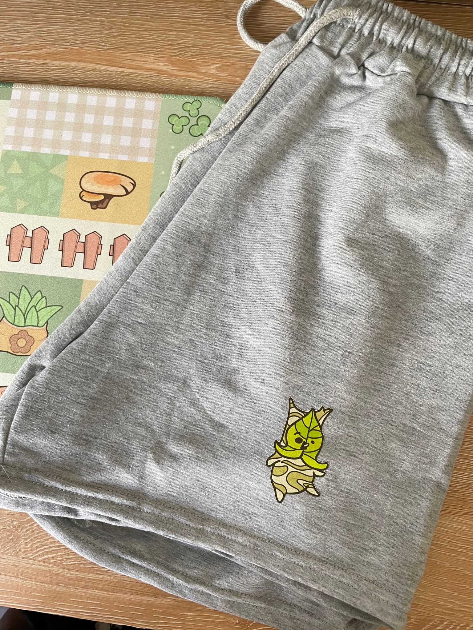 The pair of Korok Shorts OOPSIES by petimint, featuring a green leafy character with a crown on gray fabric, are laid out on a wooden surface. Next to the shorts, there is a colorful mat adorned with graphics of plants, mushrooms, and fences—perfect for any cozy gamer inspired by Stardew Valley.