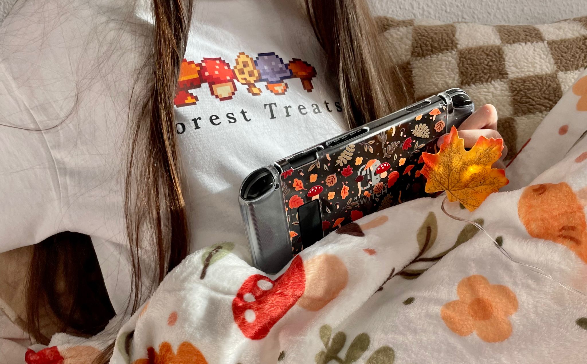 A cozy gamer with long hair holds a Nintendo Switch adorned with a mushroom-themed cover. They are wearing a white shirt with the text "Forest Treats" and a mushroom graphic. Covered by a cozy blanket with an autumn design, they also hold a small artificial maple leaf.