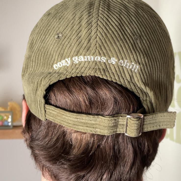 Rear view of a person with brown hair wearing an olive Cozy Gamer Club cap from petimint. The cotton cap features an adjustable strap with a metal buckle and white embroidered text that reads, "cosy games & chill." In the background, there's a shelf adorned with small framed pictures and a partially visible plant.