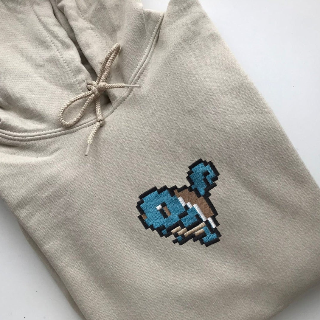 A neatly folded Squírtle Hoodie Gamer Apparel by petimint in beige features a light brown drawstring. The center of the hoodie is adorned with a pixel art design of a small blue bird, perfect for the cozygamer. The pixel art incorporates shades of blue and brown, giving the bird a retro video game look.