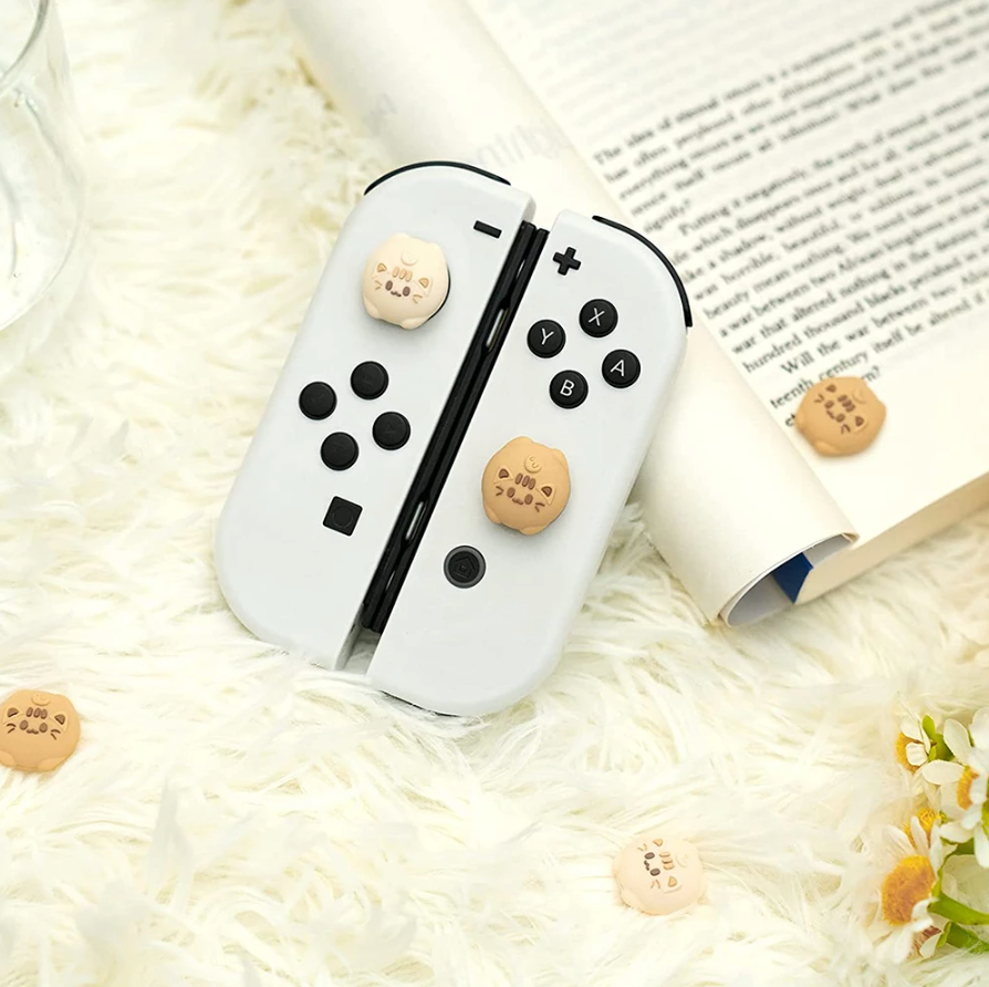 Two Nintendo Switch controllers with petimint's Soft Cats Thumb Grips rest on a white, fluffy surface. An open book and small cat-shaped thumb grips are scattered around, creating a cozy gamer scene. A part of a flower and a glass of water are also visible, completing the relaxed ambiance.