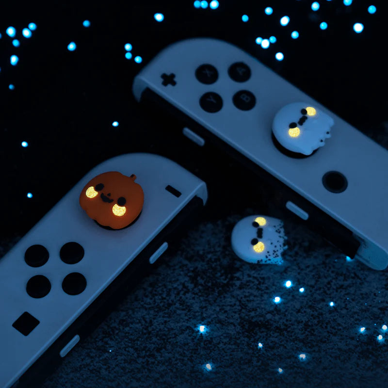 A close-up of two Nintendo Switch gaming controllers showcases petimint's Halloween Thumb Grips, which glow in the dark. One grip displays a smiling ghost, while the other sports a cheerful pumpkin. The scene is set against a dark backdrop with scattered blue lights, crafting a playful and spooky vibe ideal for late-night gaming.